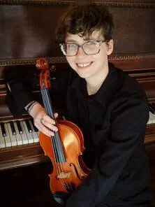 Young person with glasses smiling, holding a violin, sitting at a piano.