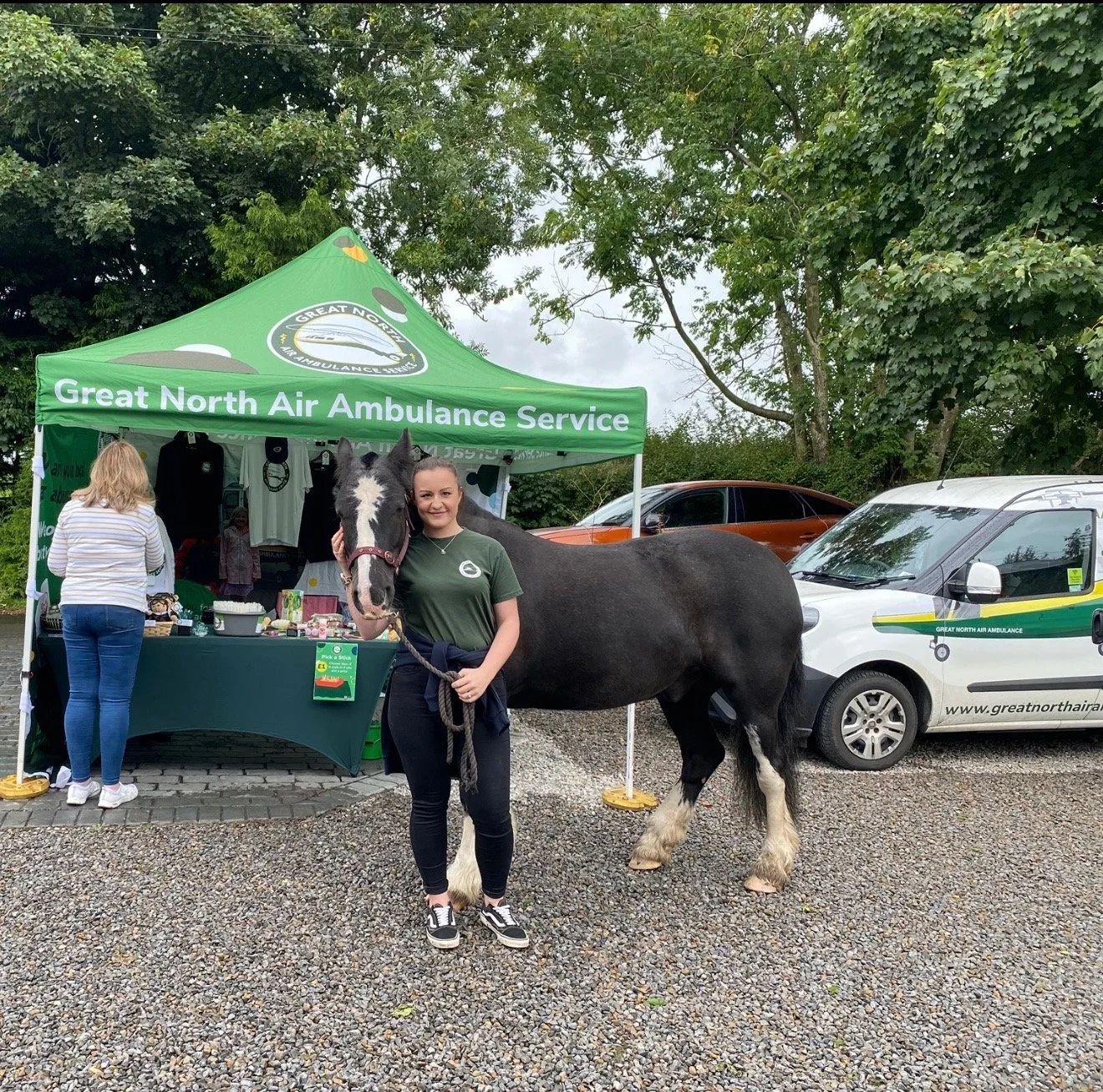A woman with a black and white horse standing in front of a green tent with the text 'Great North Air Ambulance Service.' Behind them, a white vehicle with yellow and green stripes and the website 'greatnorthair' is parked. Two other women are seen near the tent, one wearing a striped top.