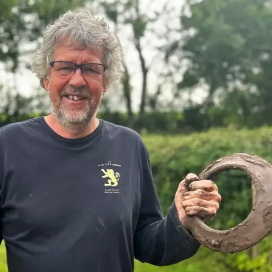 Older man with gray hair, glasses, and a beard smiling outdoors while holding a quoit