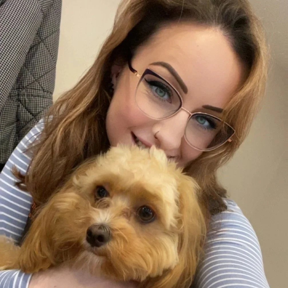 A woman with glasses and makeup hugging a small, tan, curly-haired dog.