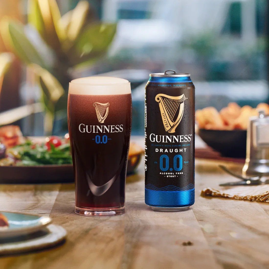 A can of Guinness alcohol-free stout and a glass filled with the same beverage on a wooden table, with a blurred background of food and plants.