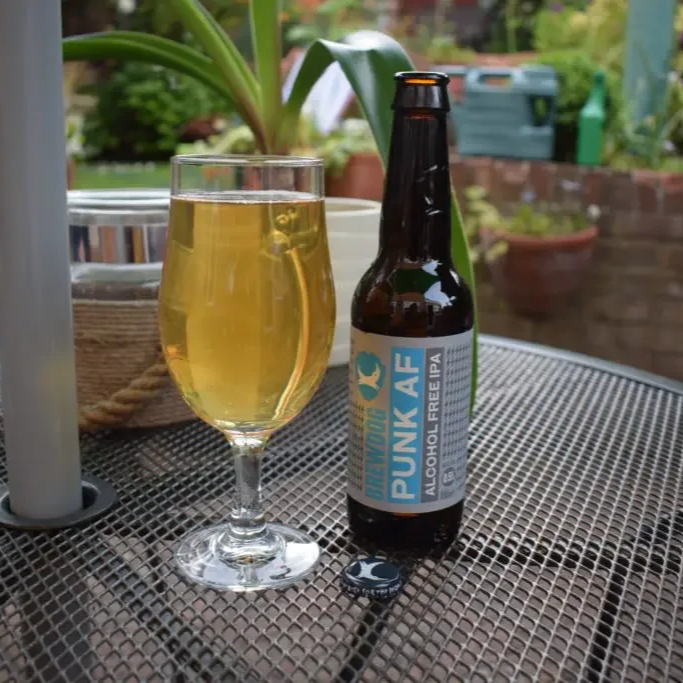 A bottle of Bravoure Punk AF alcohol-free IPA beer and a glass filled with the beer on a outdoor metal mesh table. In the background, there are potted plants and a garden setting.