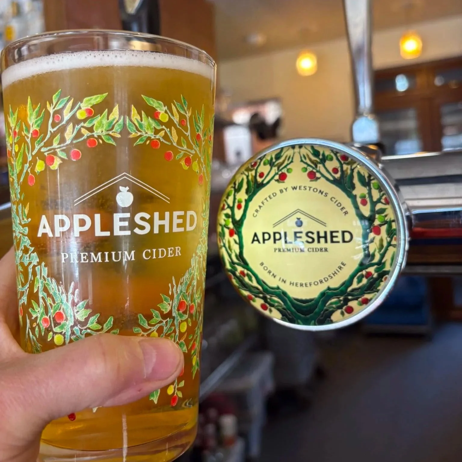 A glass of apple cider with a decorative label featuring apple trees, branded as 'Appleshed Premium Cider' and a can with the same branding in the background.