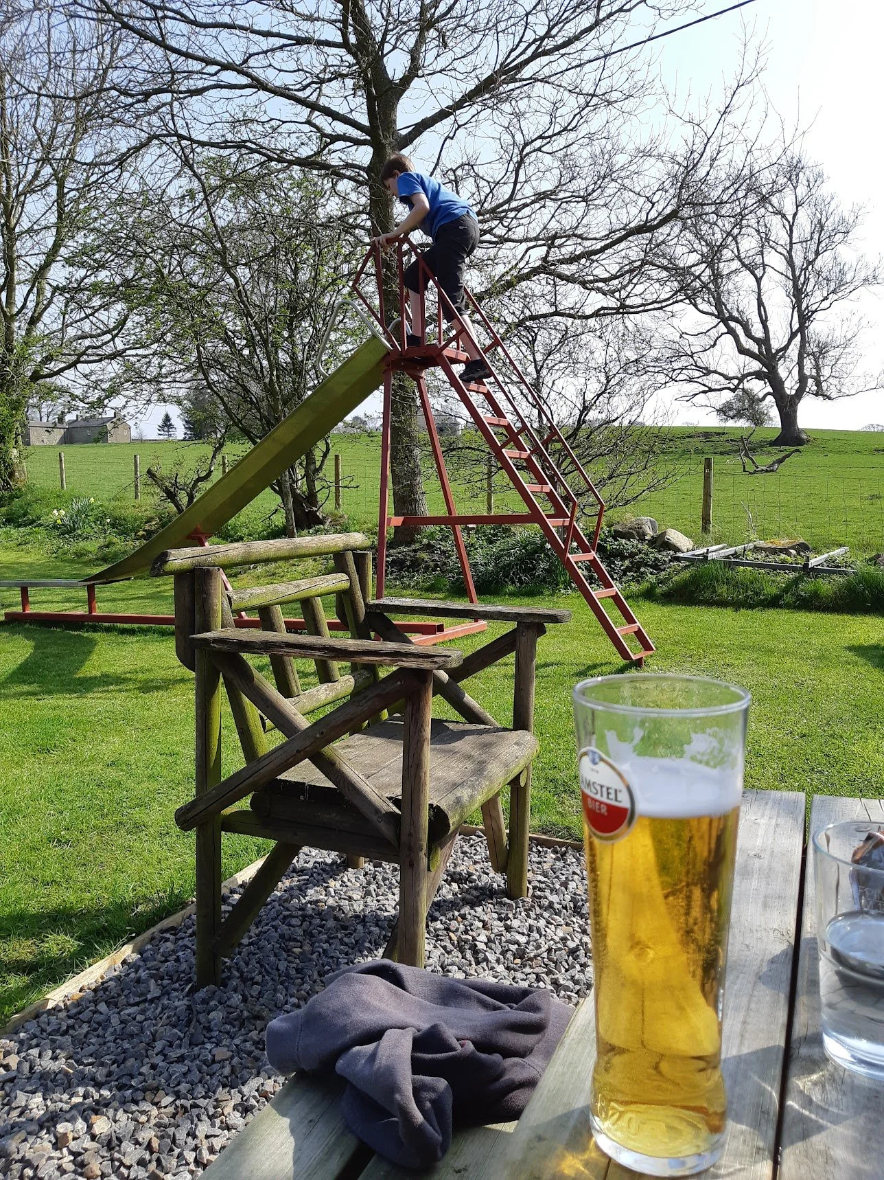 Child climbing a slide on a playground set in a backyard with green grass, trees, and a fence. A table with a glass of beer and a glass of water is in the foreground.