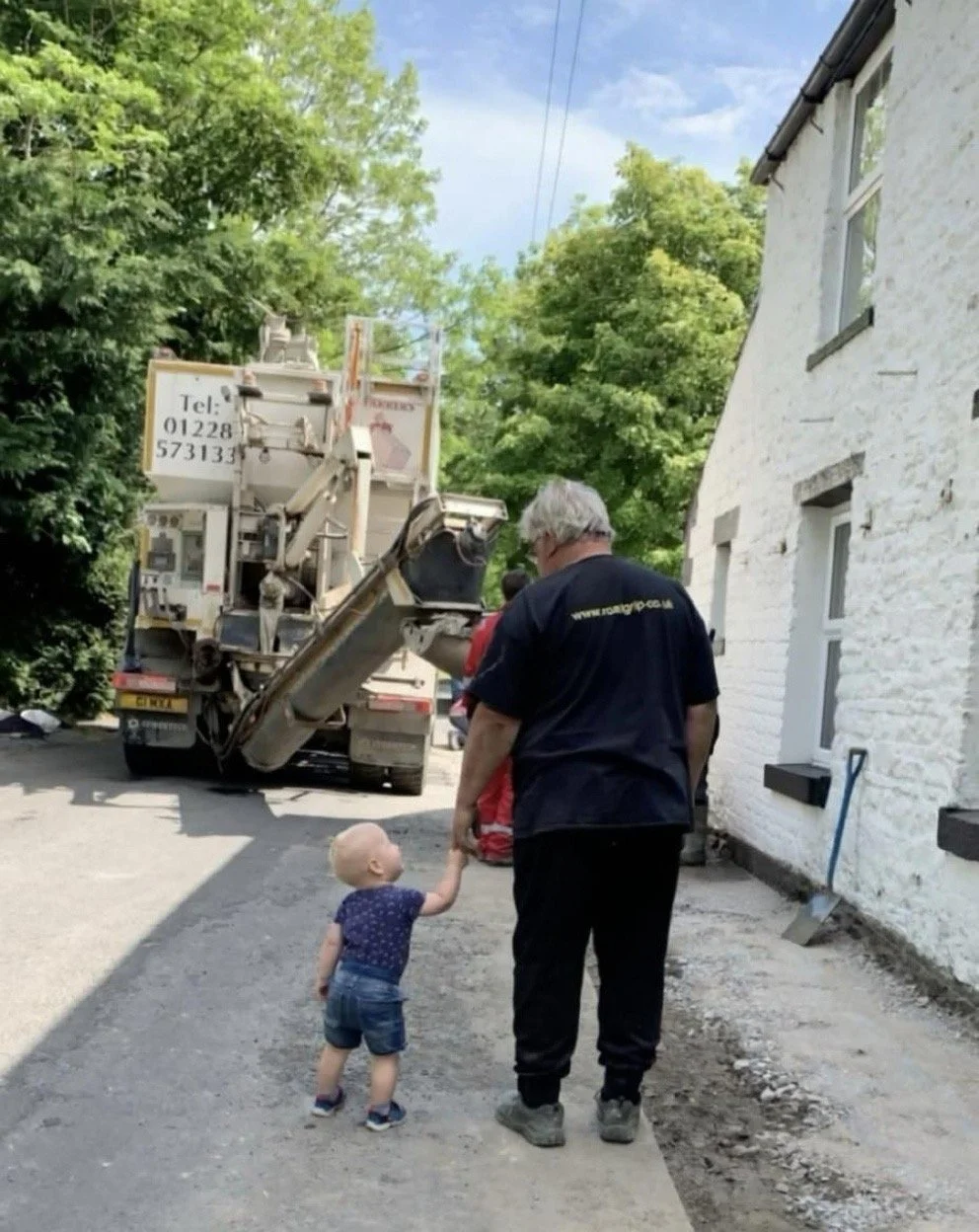 A young child holding hands with an older man on a sidewalk, near a white brick house, with a large truck parked nearby and trees in the background.