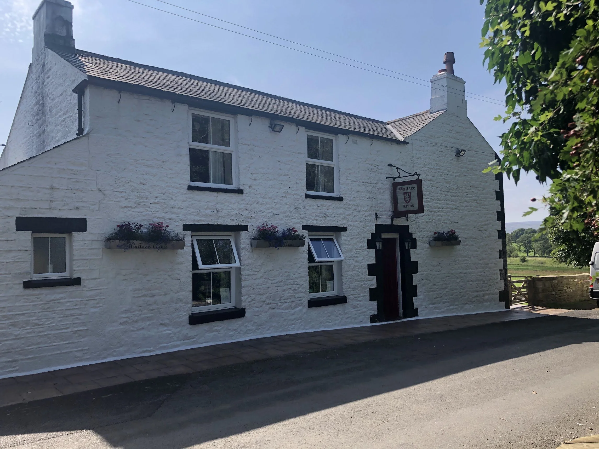 A white stone building with black accents, sign reading 'Wallace Arms,' and flower boxes on some windows.