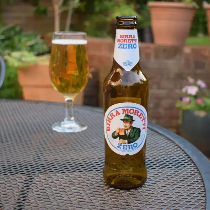 A bottle of Birra Moretti Zero beer with a label showing a man in a green suit and hat holding a beer, placed on an outdoor metal table with a glass of beer in the background and potted plants.