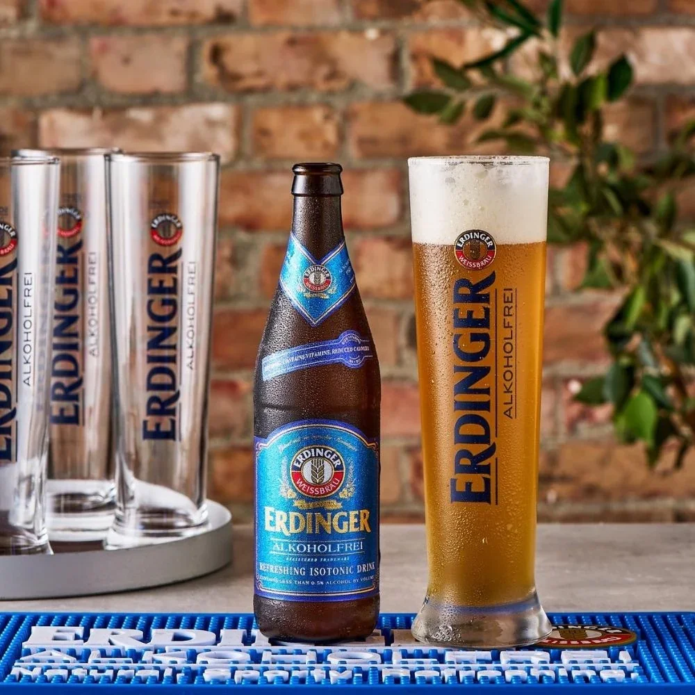 A glass and a bottle of Erdinger alcohol-free beer, with the glass filled with beer and topped with foam, set on a bar mat with other glasses in the background on a wooden table against a brick wall.