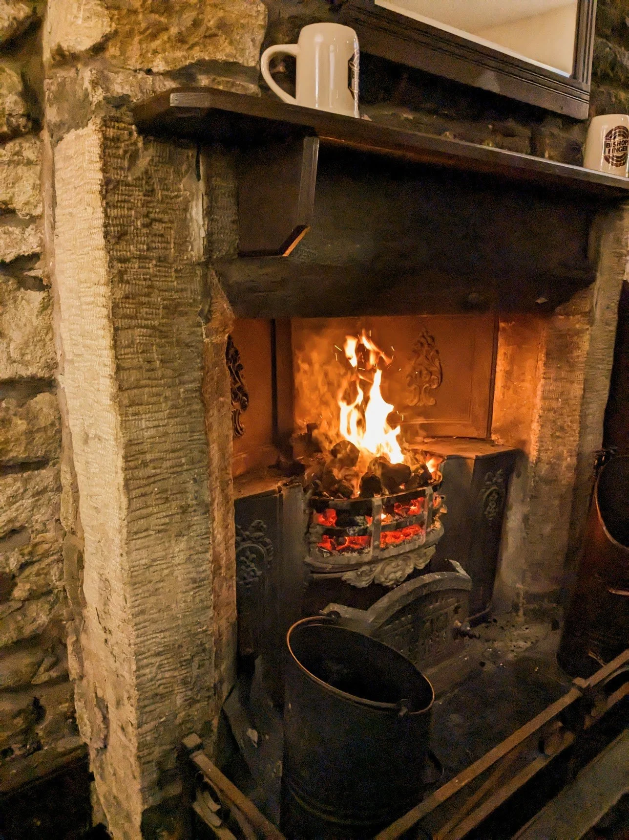 A lit fireplace with burning logs, set in a brick and stone wall. A black fire grate holds the logs, and a black bucket is placed on the hearth below. A wooden shelf above the fireplace has two white mugs and a framed mirror.