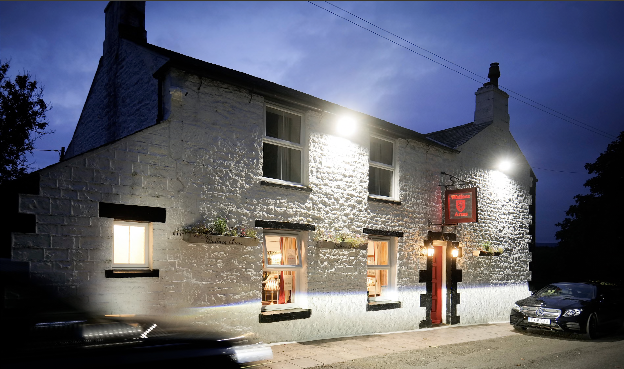 A two-story white brick building with black window frames and a red door, illuminated at night. The building has a sign reading 'Wallace Arms' and potted plants on window ledges. A black car is parked in front, and there is motion blur of passing cars on the street.