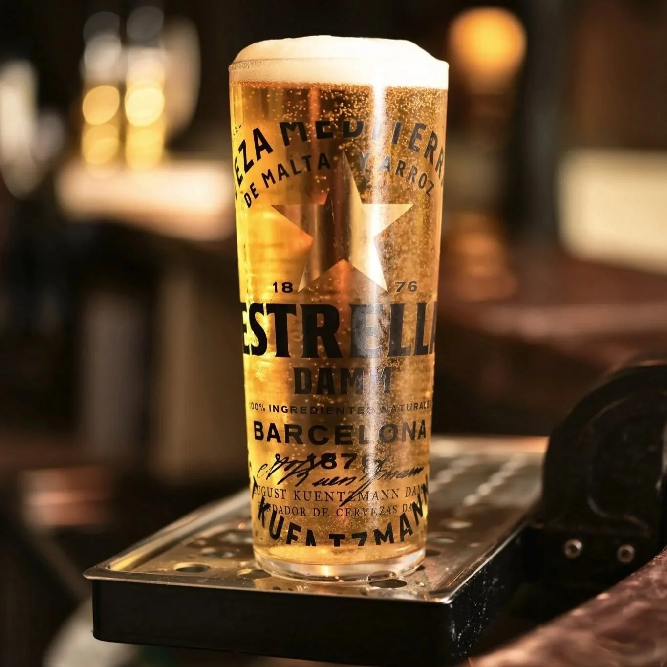 A glass of beer with foam on top, placed on a metal surface, with blurred background of bar or kitchen.