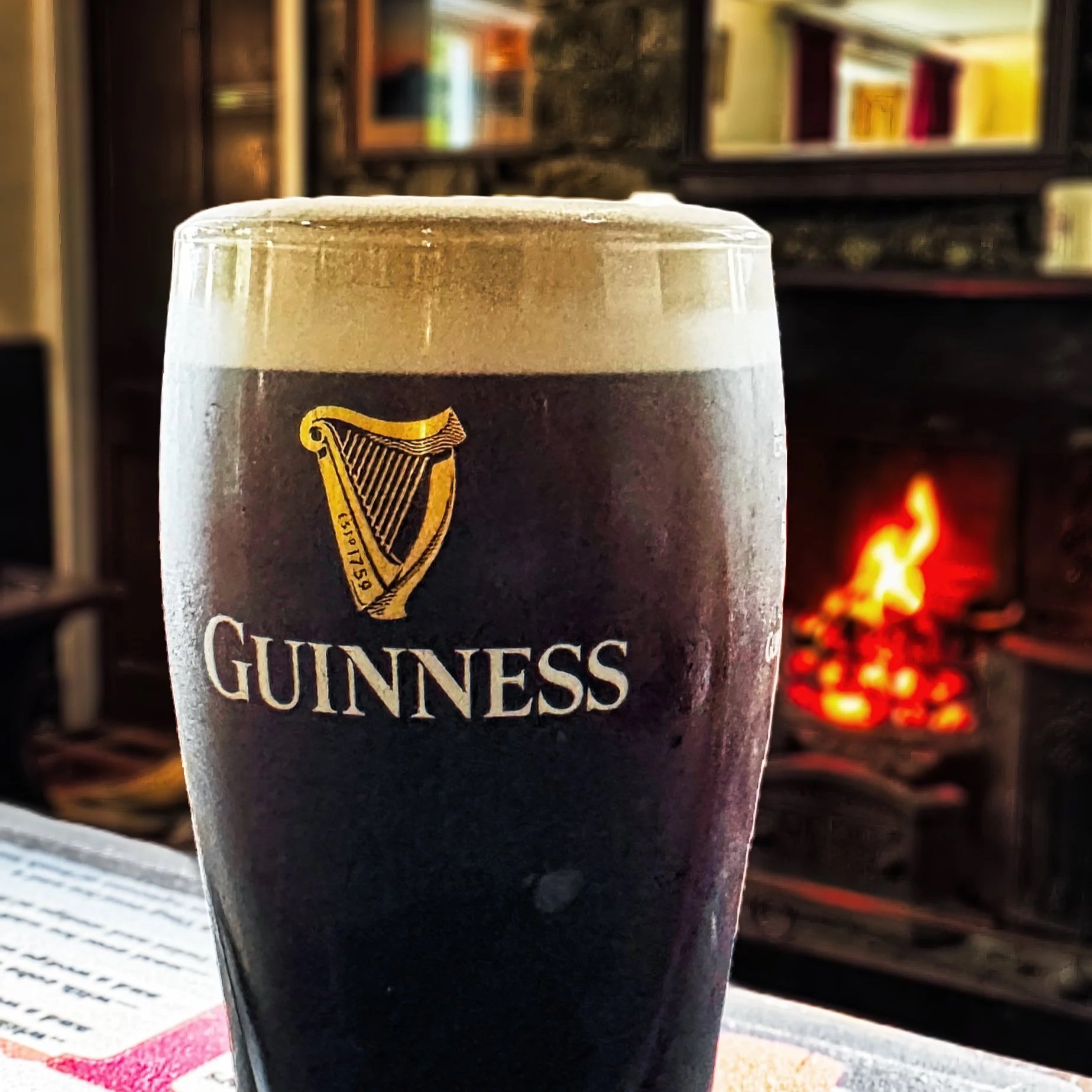 A pint of Guinness beer with a foamy head on top, placed on a table near a fireplace with a fire burning in the background.