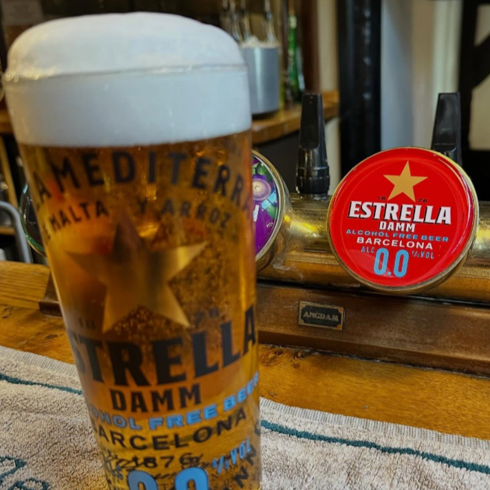 A glass of Estrella Damm beer with foam on top, placed on a wooden bar counter. In the background, there is a tap with the Estrella Damm logo and other bar equipment.