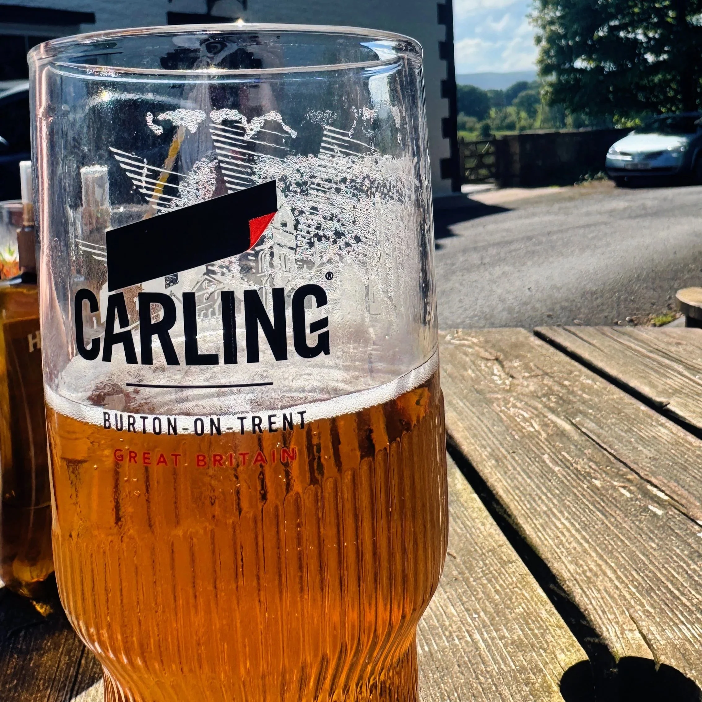 Close-up of a glass of beer on a wooden table outdoors, with a parking lot, cars, trees, and blue sky in the background.