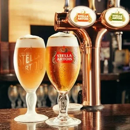 Two glasses of Stella Artois beer on a bar counter with beer taps in the background.