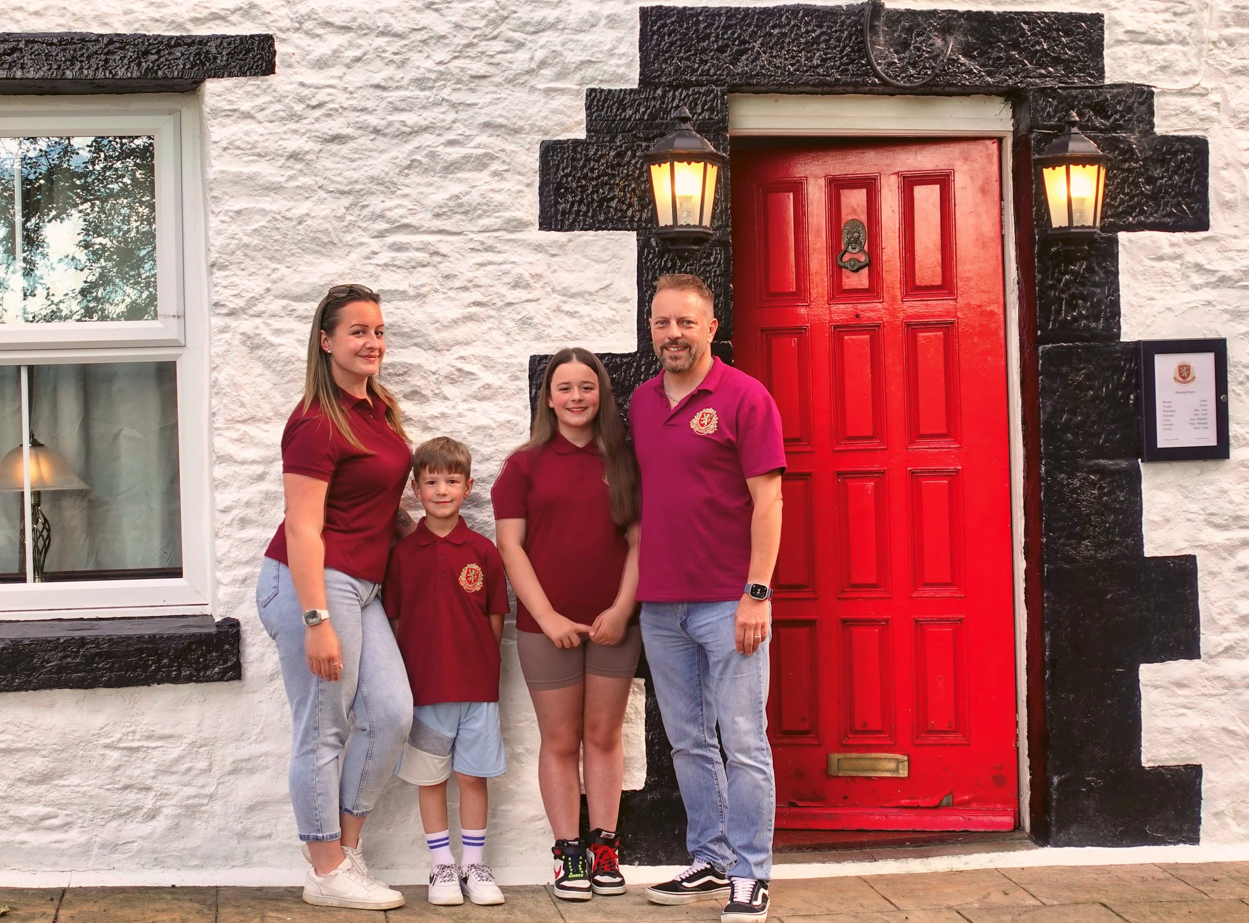 A family of four stands in front of a white house with black trim and a red door, smiling at the camera. The woman and girl are wearing matching maroon shirts with a crest, while the boy and man are dressed in similar maroon polo shirts. The woman has long brown hair and is wearing jeans, the girl has long dark hair, the boy has short hair, and the man has short hair and a beard. Two black lantern-style lights flank the red door, which has a lion head door knocker and a mail slot below.