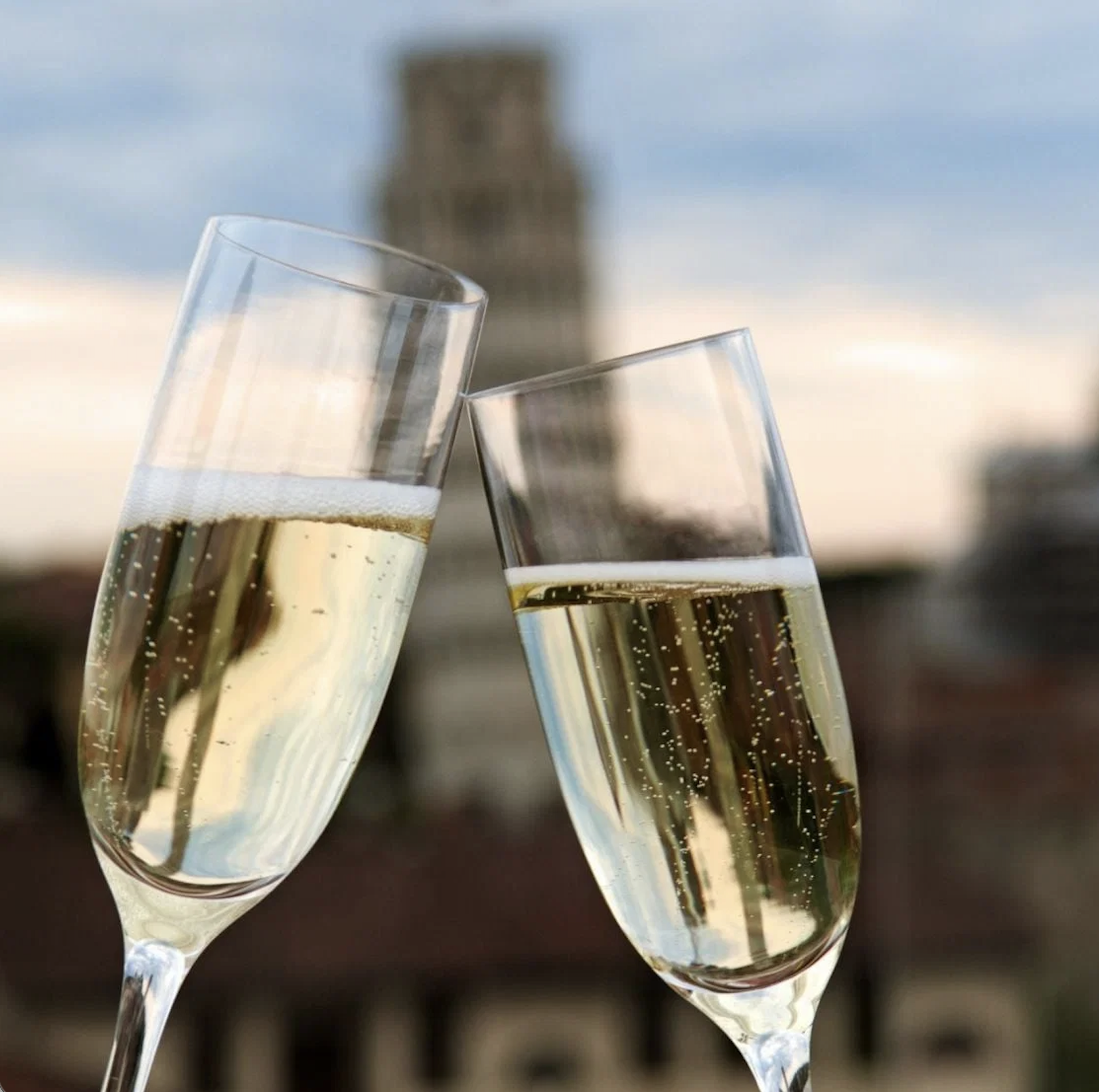 Two champagne glasses being clinked together in a toast, with the Piza Tower blurred in the background.