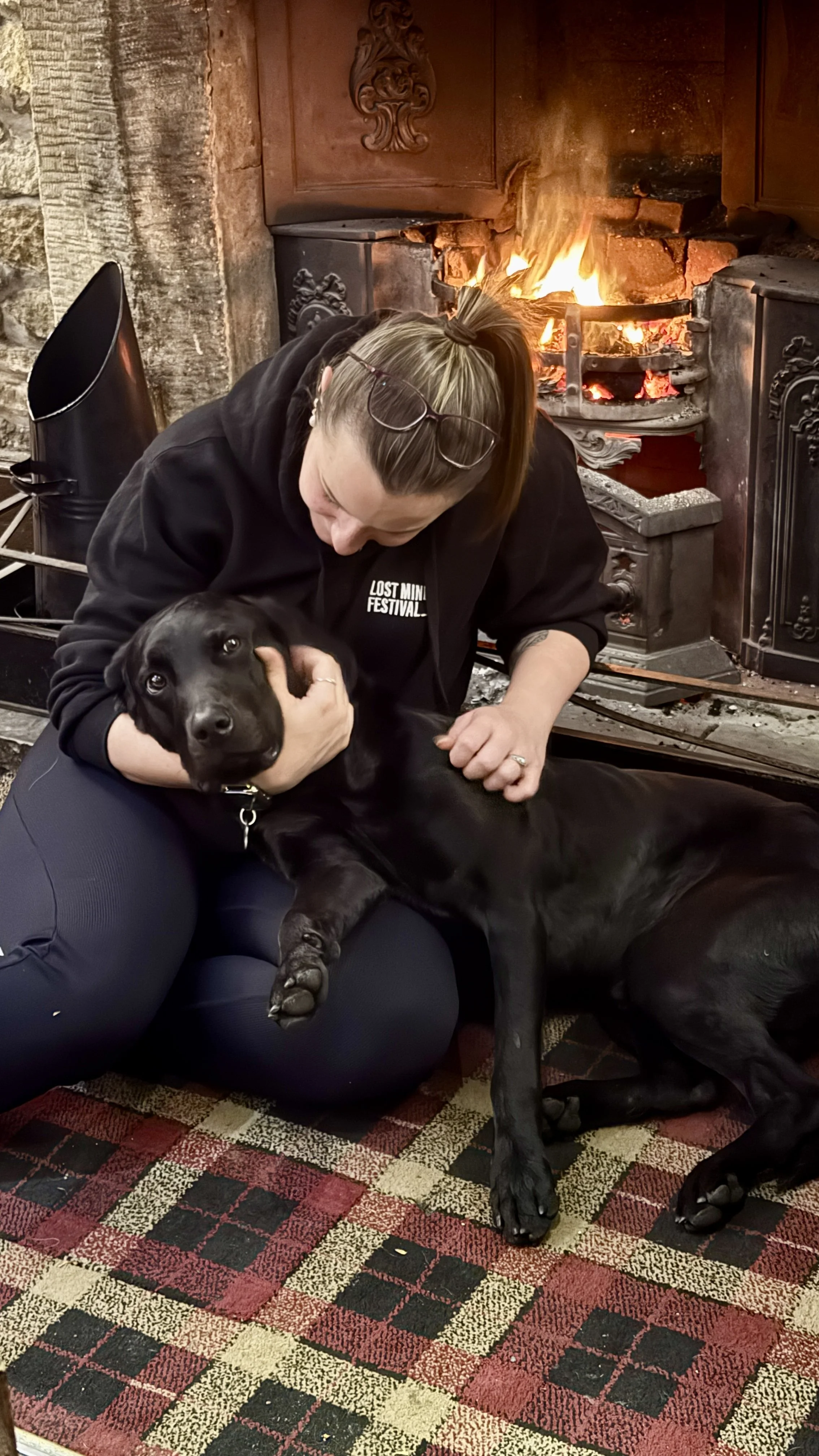 A woman with glasses on her head and earrings hugs a black Labrador retriever puppy while sitting in front of a fireplace with a fire burning inside.
