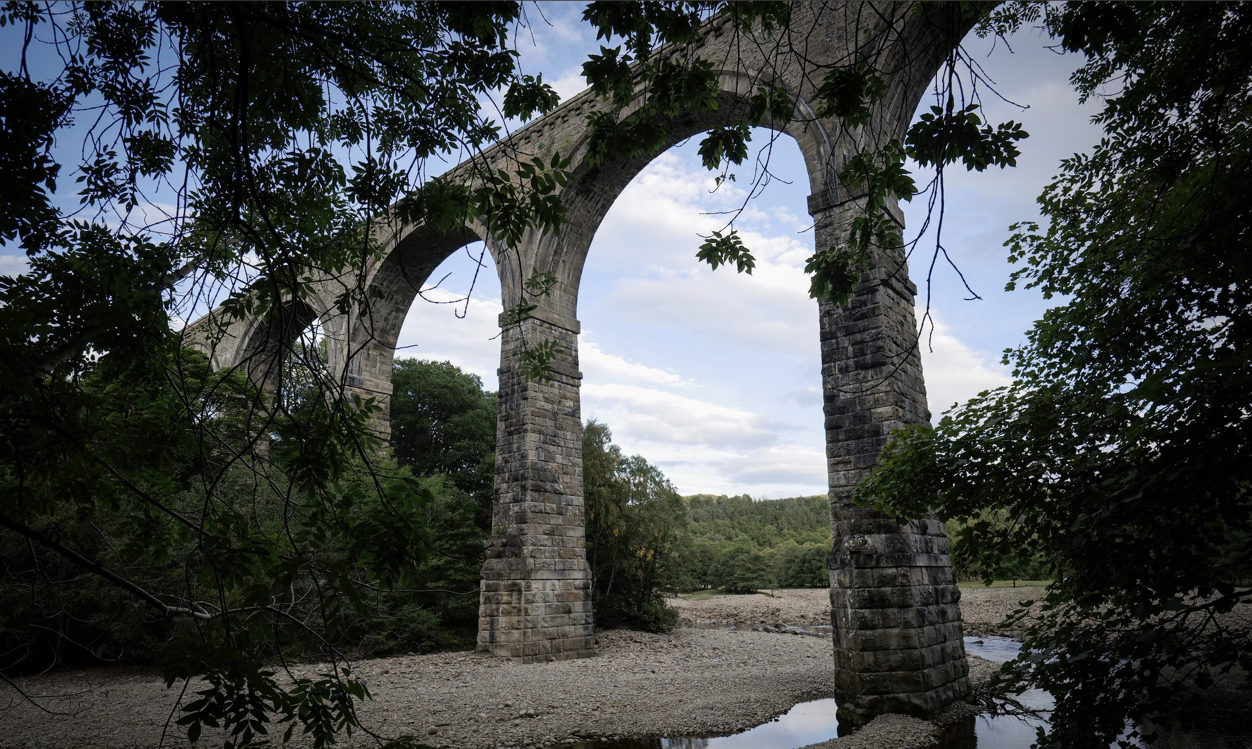 Stone arch bridge over a shallow river, surrounded by trees with a cloudy sky in the background.