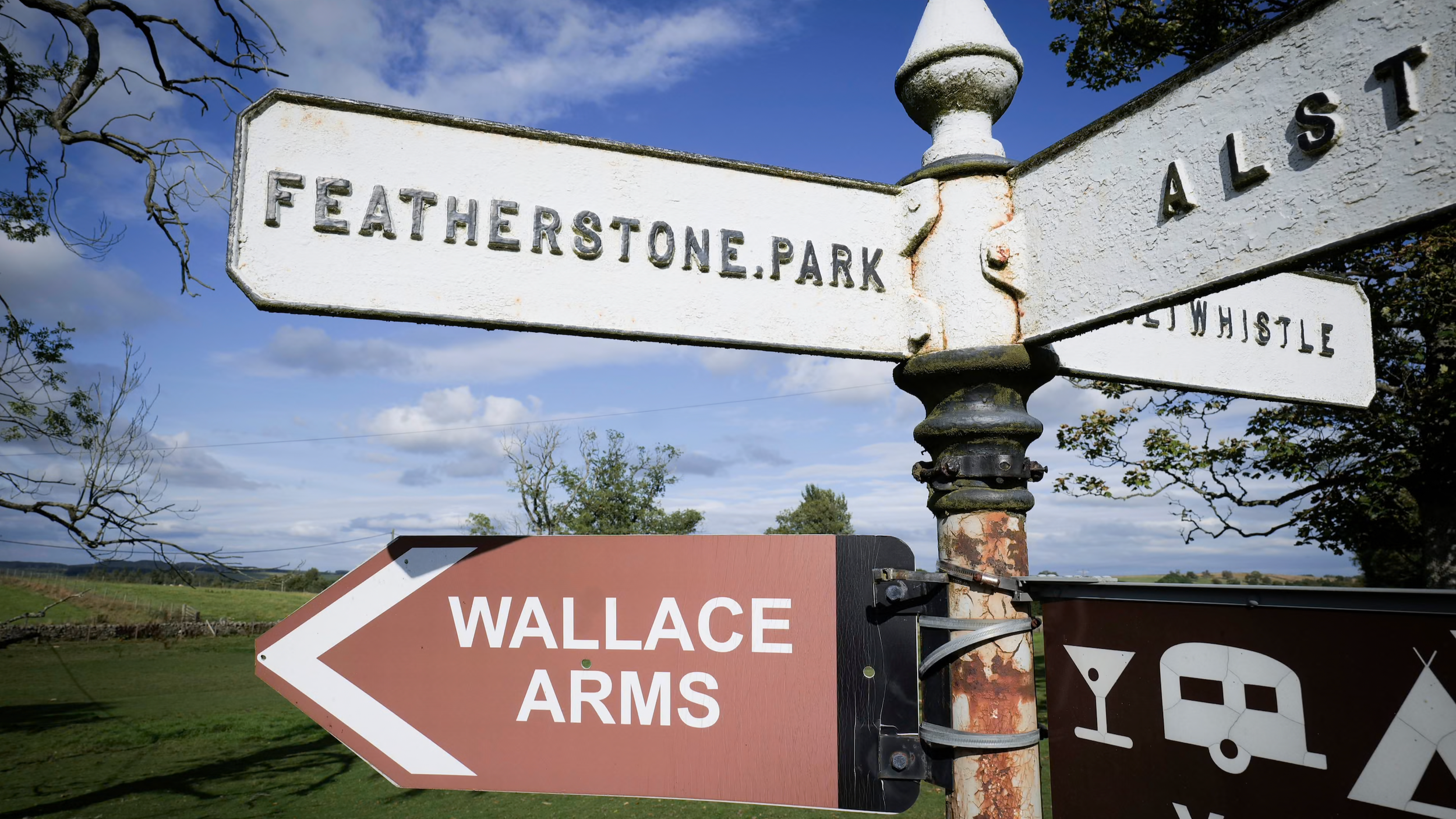 A street sign pointing to Featherstone Park, a sign pointing to Wallance Arms, and a sign indicating a pub or bar, with a rural landscape and blue sky in the background.