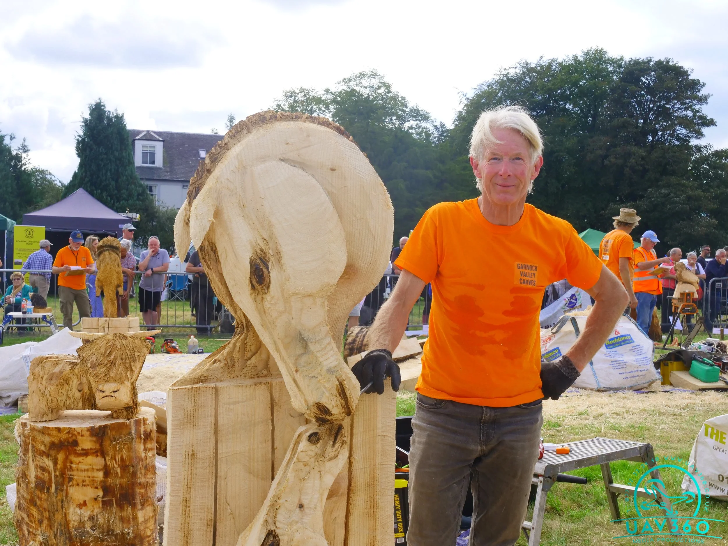 A man with white hair and a smiling expression is standing outdoors at a woodworking event, wearing an orange T-shirt and black gloves, next to a large, partially carved wooden sculpture of an animal head, with people and tents visible in the backgro