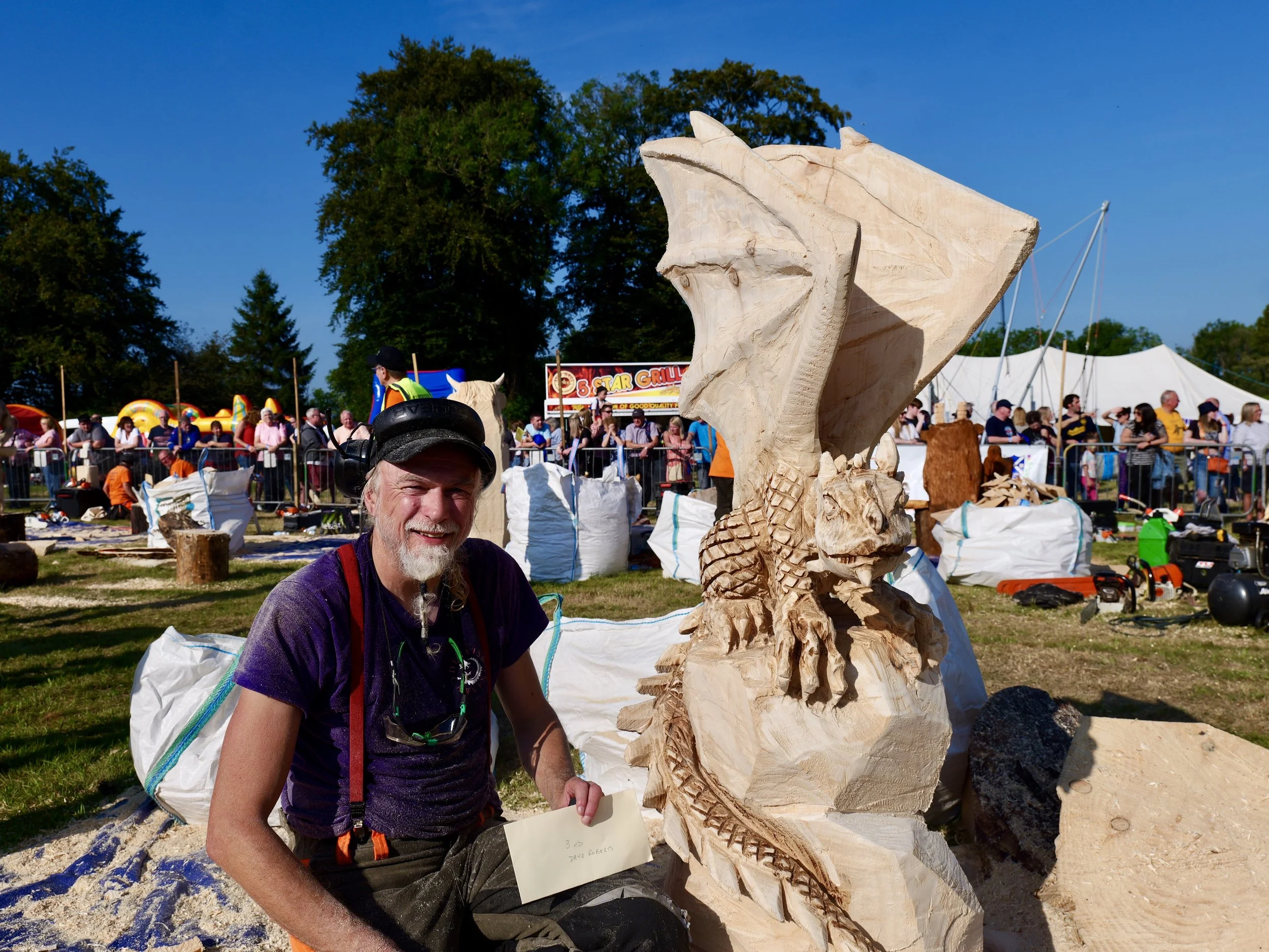 A man with a beard and hat smiling next to a large wooden sculpture of a dragon with wings and a lion's head at an outdoor event with a crowd of people and tents in the background.