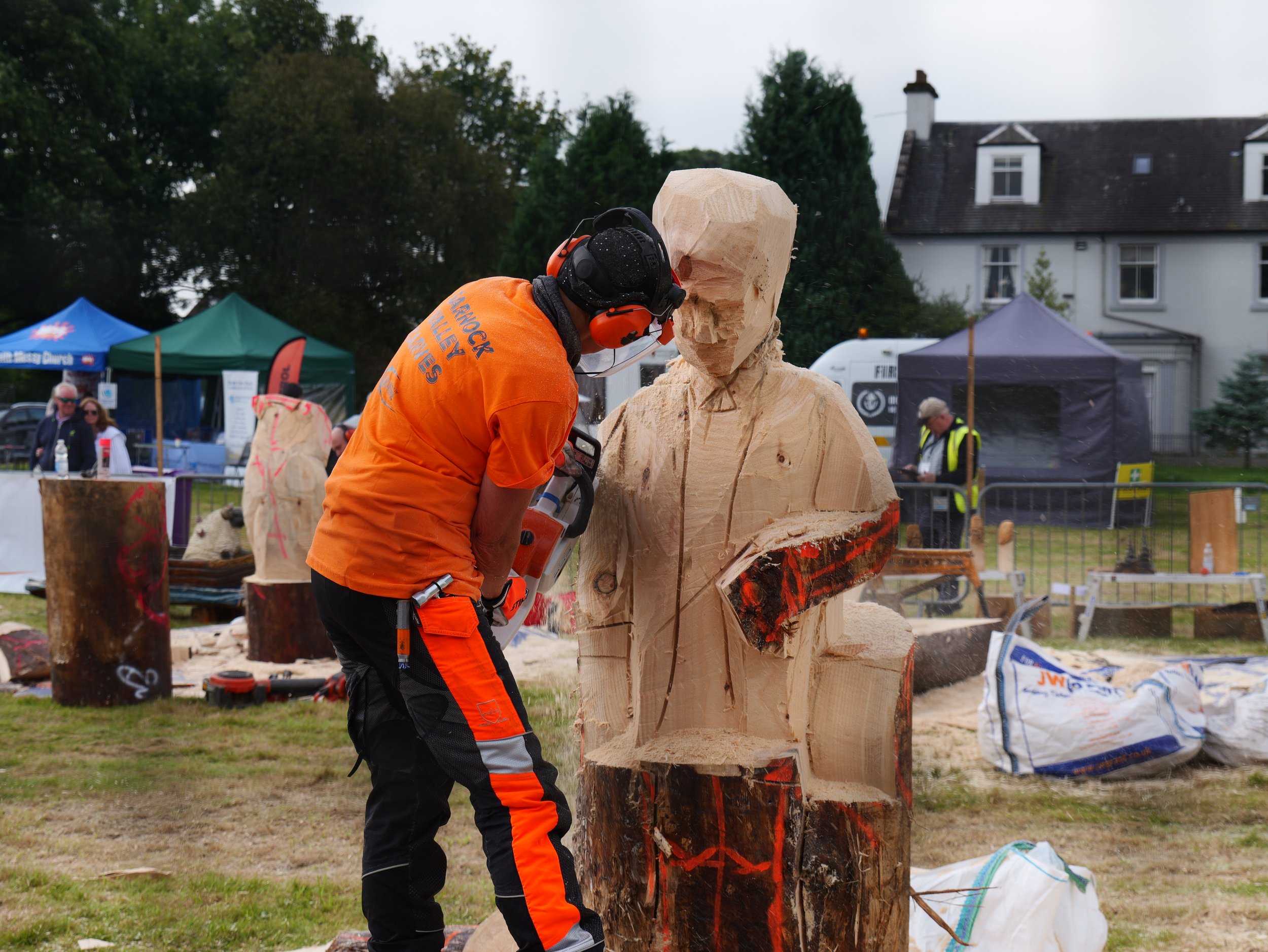 A person wearing safety gear and an orange shirt using a chainsaw to carve a large wooden sculpture of a man in an outdoor event area.