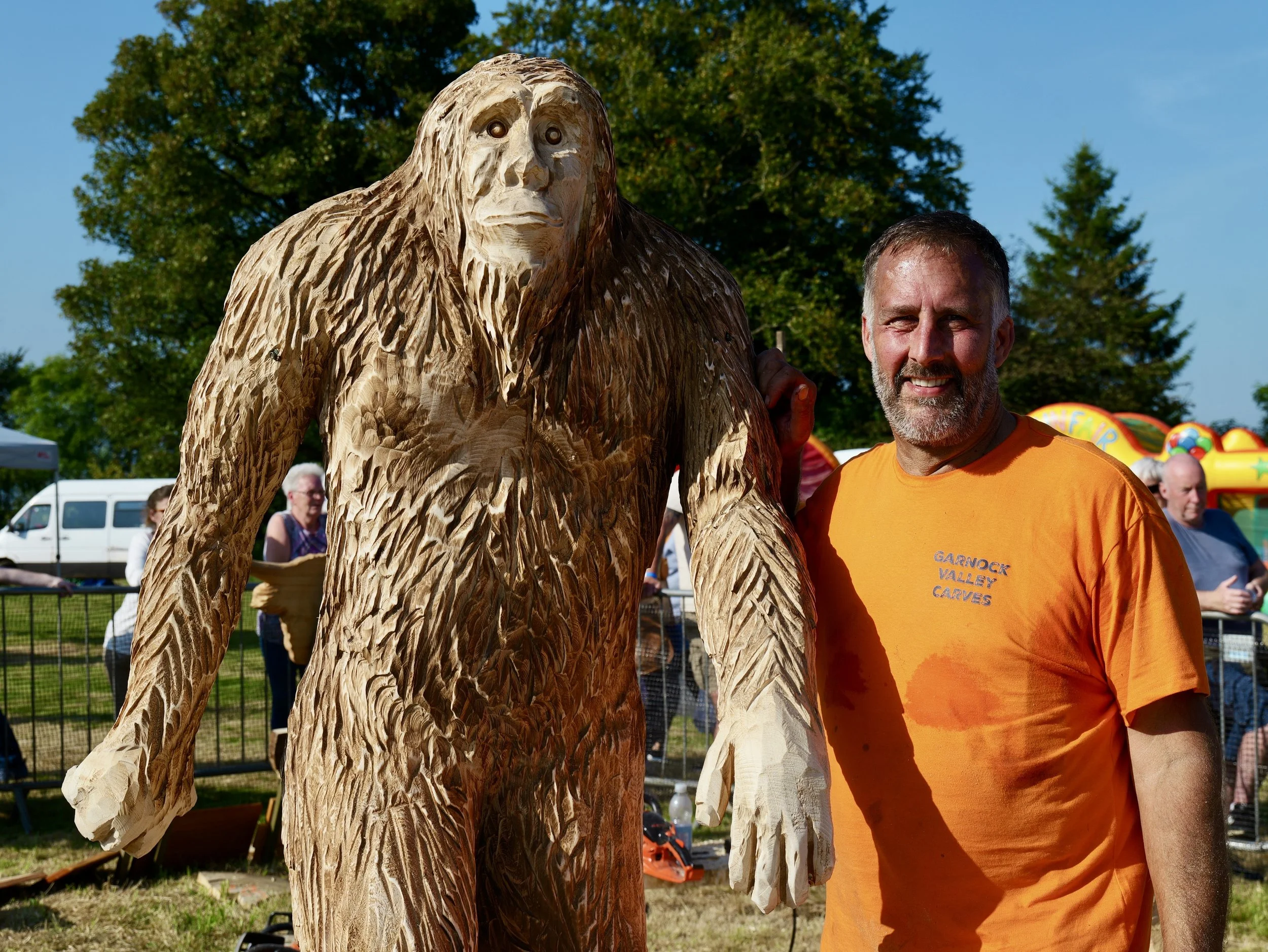 A man in an orange shirt standing next to a large wooden gorilla sculpture at an outdoor event, with people and trees in the background.