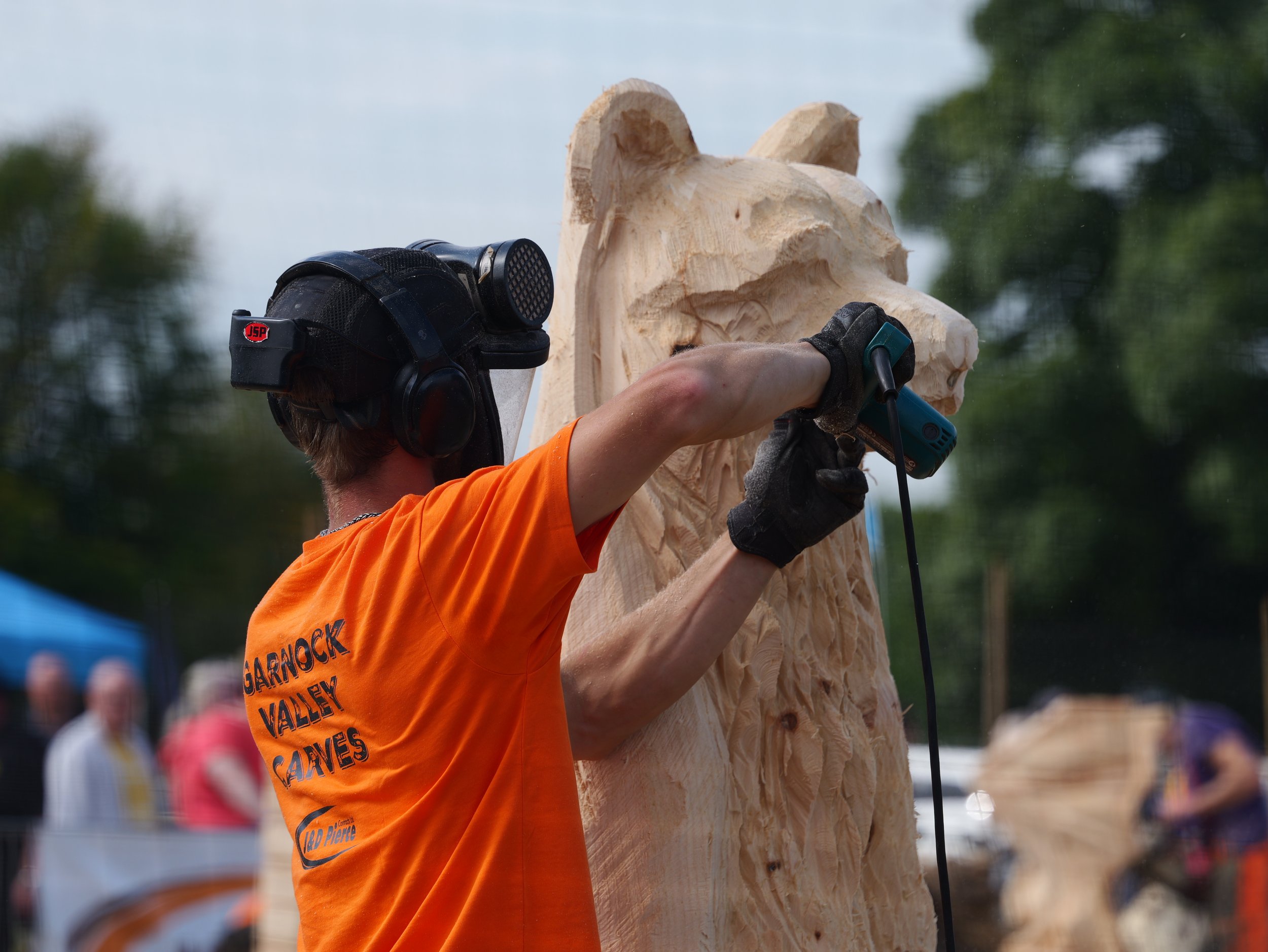 A man carving a large wood sculpture of an animal's head, wearing safety gear, during an outdoor event.