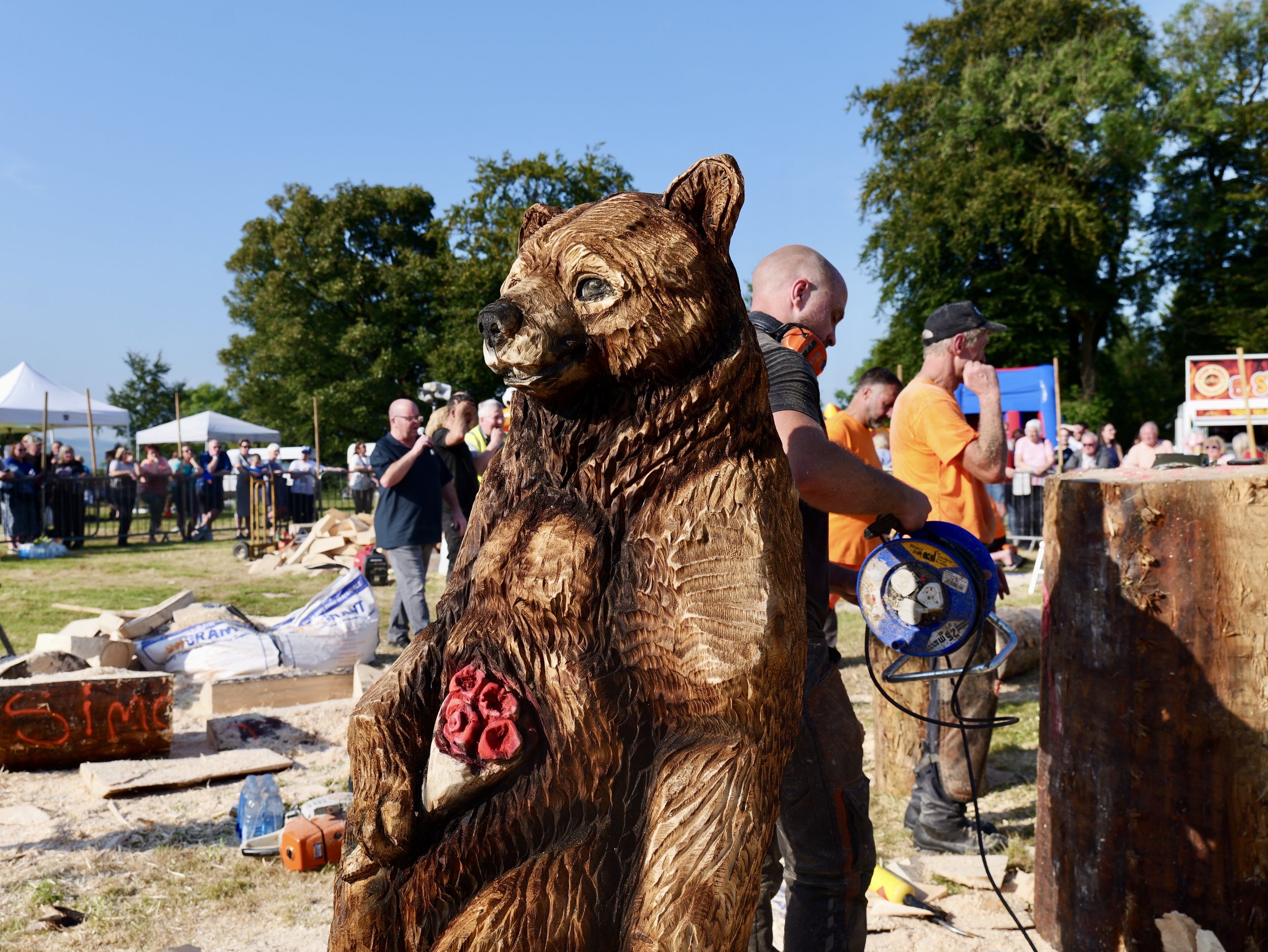 A large wooden bear sculpture in the foreground at an outdoor event, with people and tents in the background.