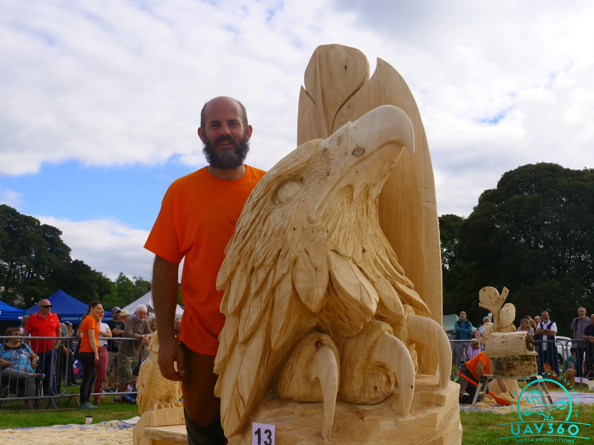 A man with a beard in an orange shirt stands next to a large wooden sculpture of an eagle with a mountain background at an outdoor event with people and tents.