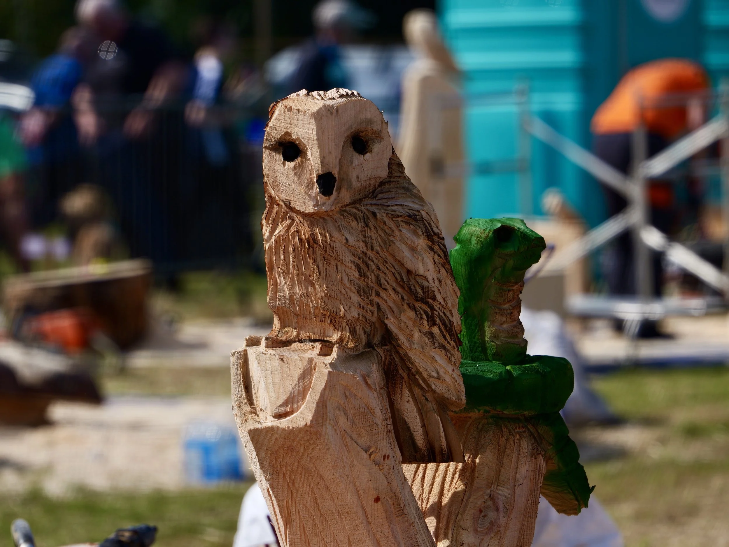 Wooden carving of an owl at an outdoor craft fair, with blurred background of people and booths.