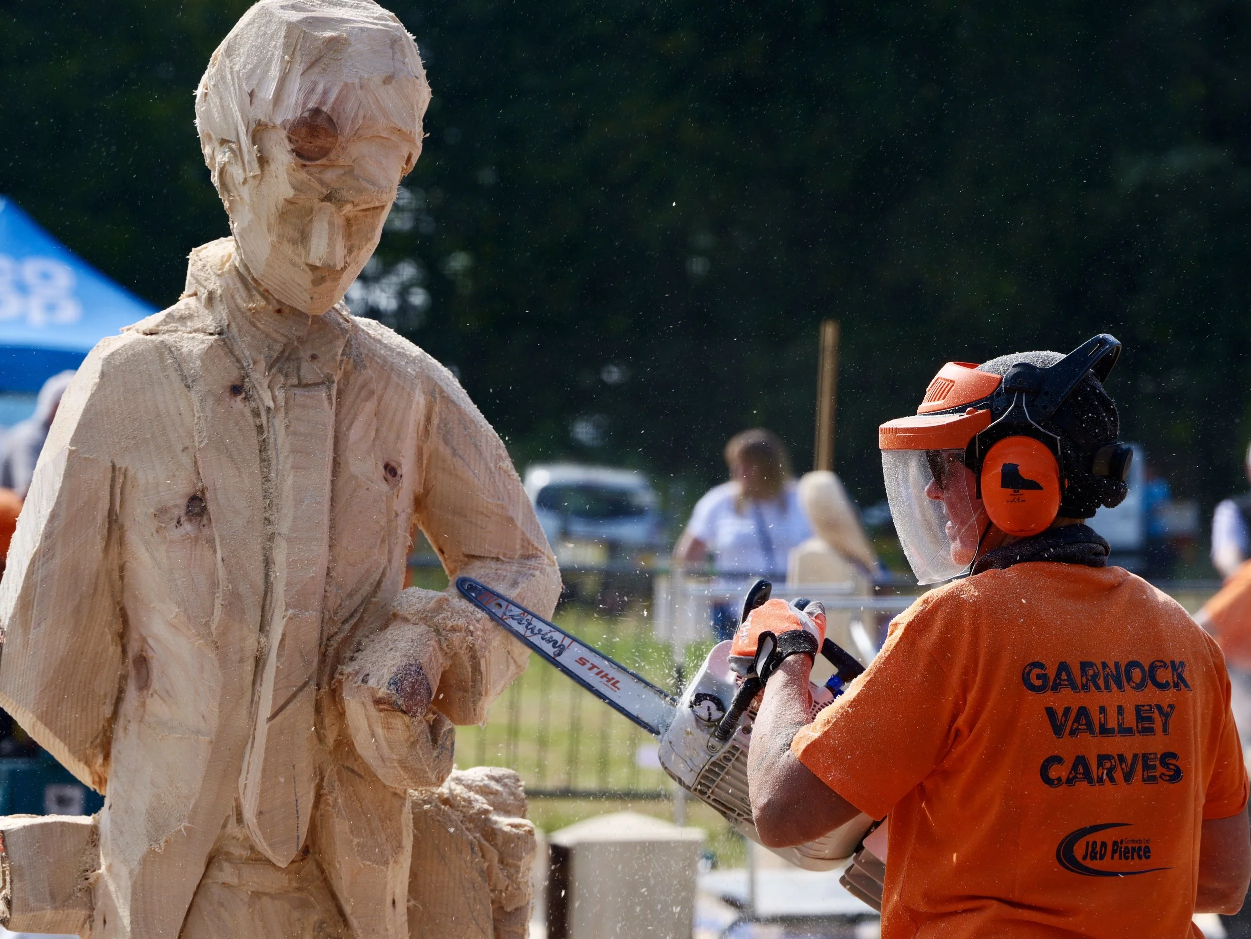A person in orange protective gear and a helmet uses a chainsaw to carve a large wooden sculpture of a man against a background of trees and people in an outdoor setting.