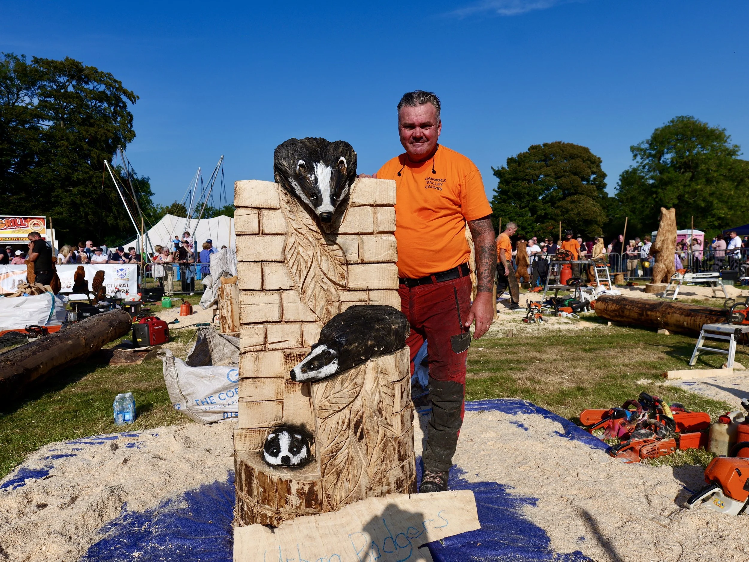 Man standing next to a carved wood sculpture of a weasel's head with a large wooden block featuring additional weasel faces, at an outdoor event with many people and art displays in the background.