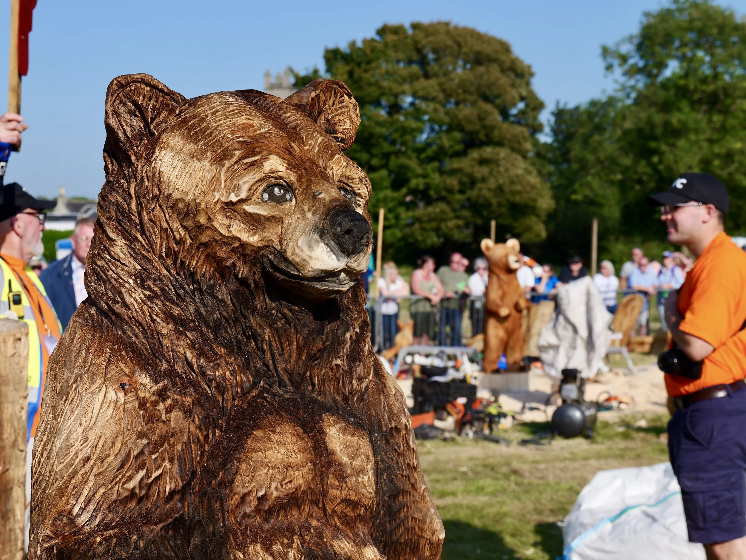 A detailed wooden sculpture of a bear at an outdoor event, with people and trees in the background.
