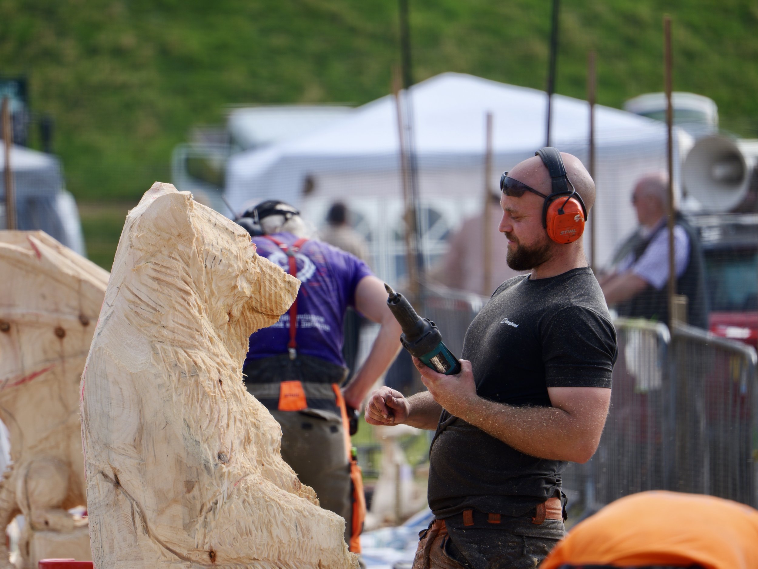 Man wearing red ear protection and black t-shirt working on a wood carving at an outdoor event.