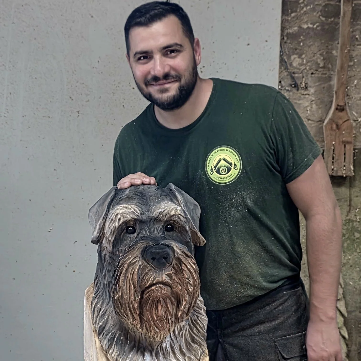 A man with dark hair and a beard, wearing a green shirt with a logo, stands inside next to a detailed carved wooden sculpture of a dog's head.