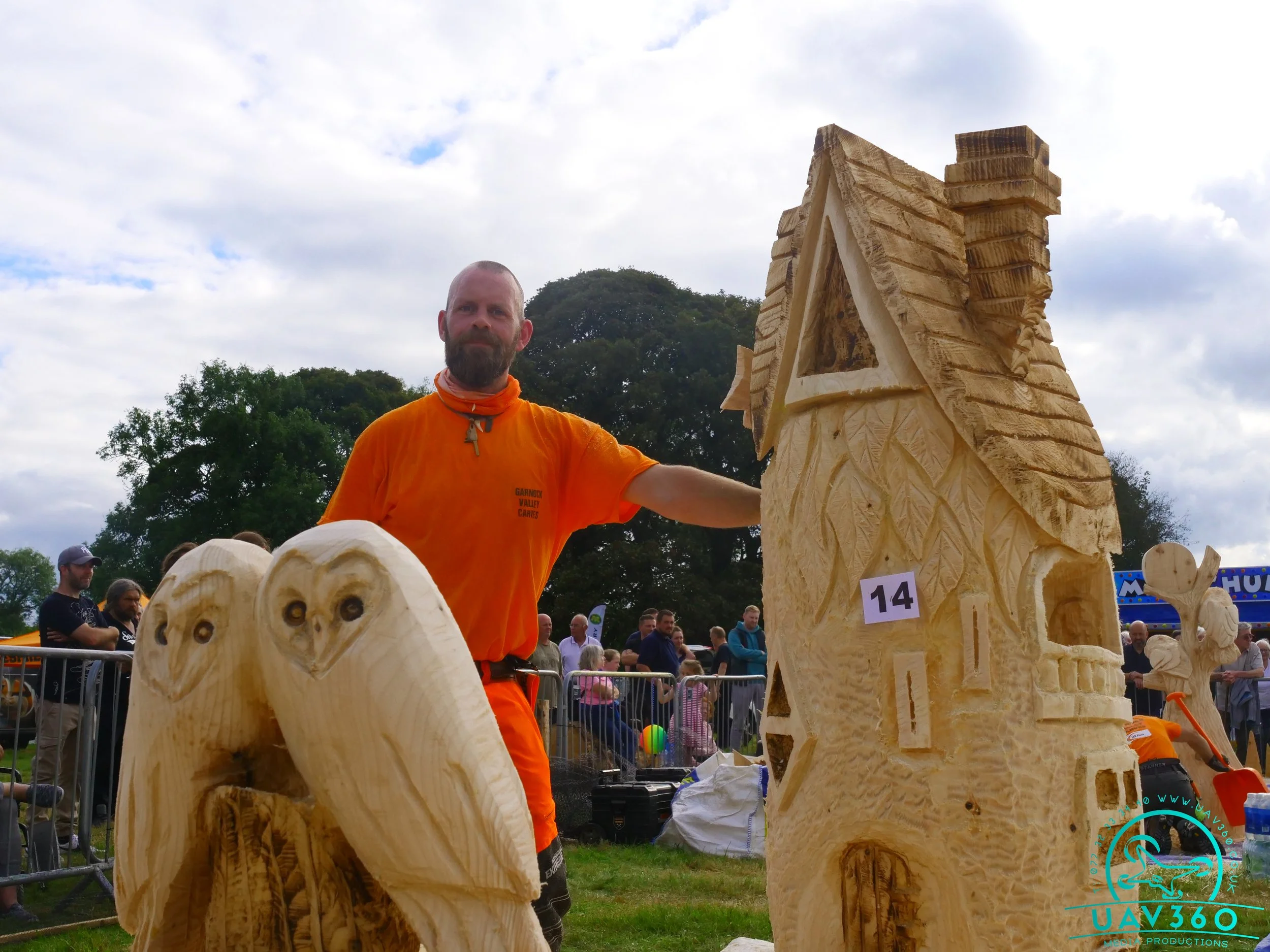 A man with a beard wearing an orange t-shirt and neck gaiter stands outdoors at an event, showcasing carved wooden sculptures of an owl and a house. There are spectators in the background, some behind barriers, with trees and a cloudy sky overhead.