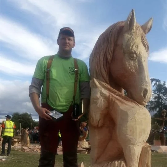 A man wearing a green shirt and a cap standing next to a large wooden horse sculpture outdoors during daytime.