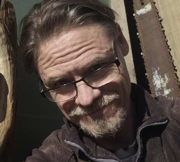 Close-up of a man with glasses, a gray beard, smiling, wearing a dark jacket in sunlight with wooden and metal background.