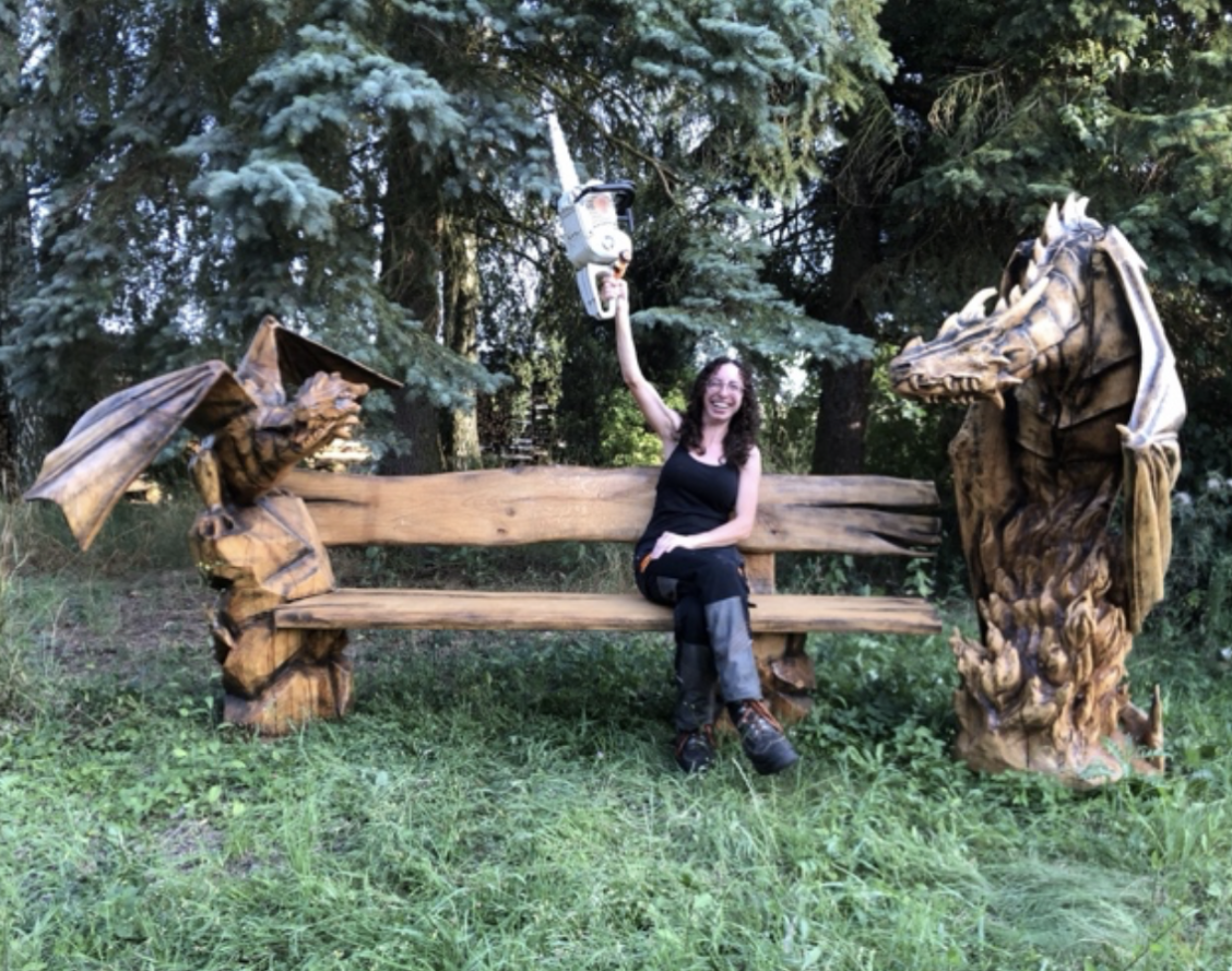 A woman sitting on a wooden bench with dragon carved armrests and backrest, smiling, outdoors among trees, holding a speaker overhead.