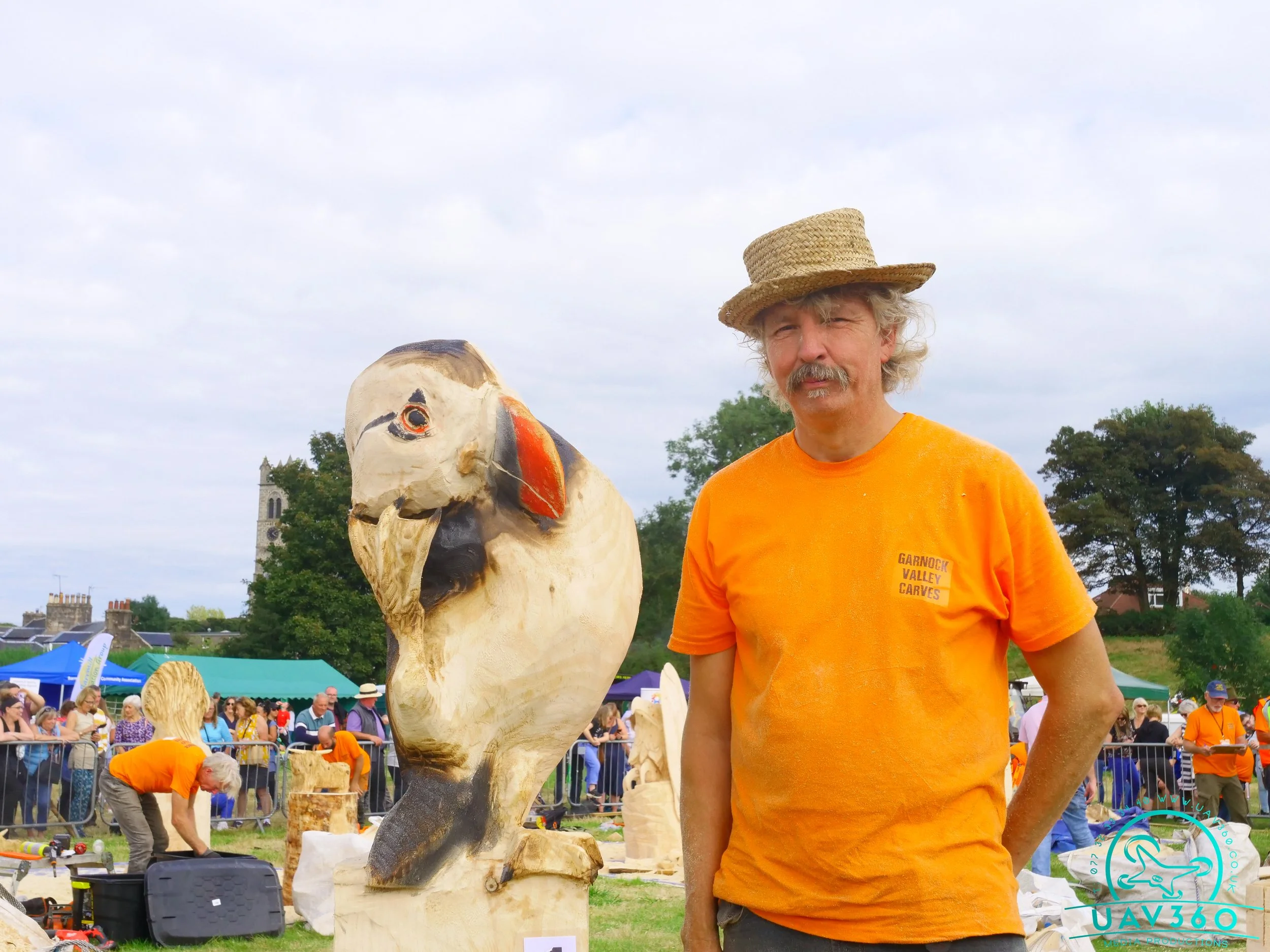 A man with gray hair and a mustache wearing an orange t-shirt and straw hat standing next to a carved wooden bird sculpture at an outdoor event.