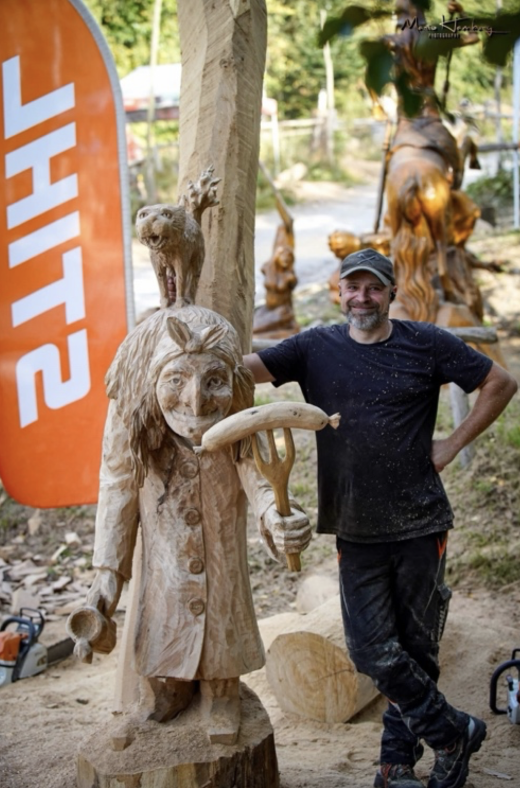 A man posing next to a wooden sculpture of a woman with a llama hat and holding a fork, and a carved lion and other animal figures in the background at an outdoor wood sculpture event.