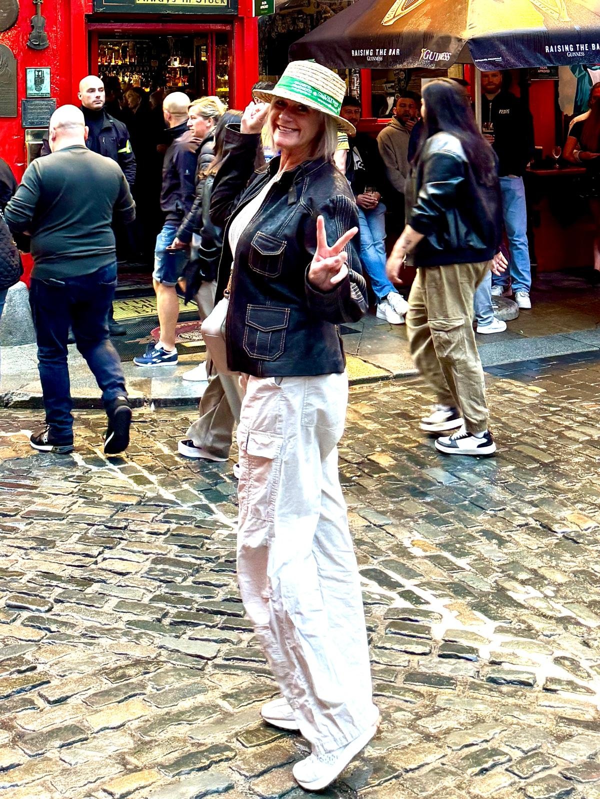 A woman in a black denim jacket and white cargo pants posing on a cobblestone street, wearing a striped straw hat and making a peace sign. The street is busy with people outside a bar or pub.
