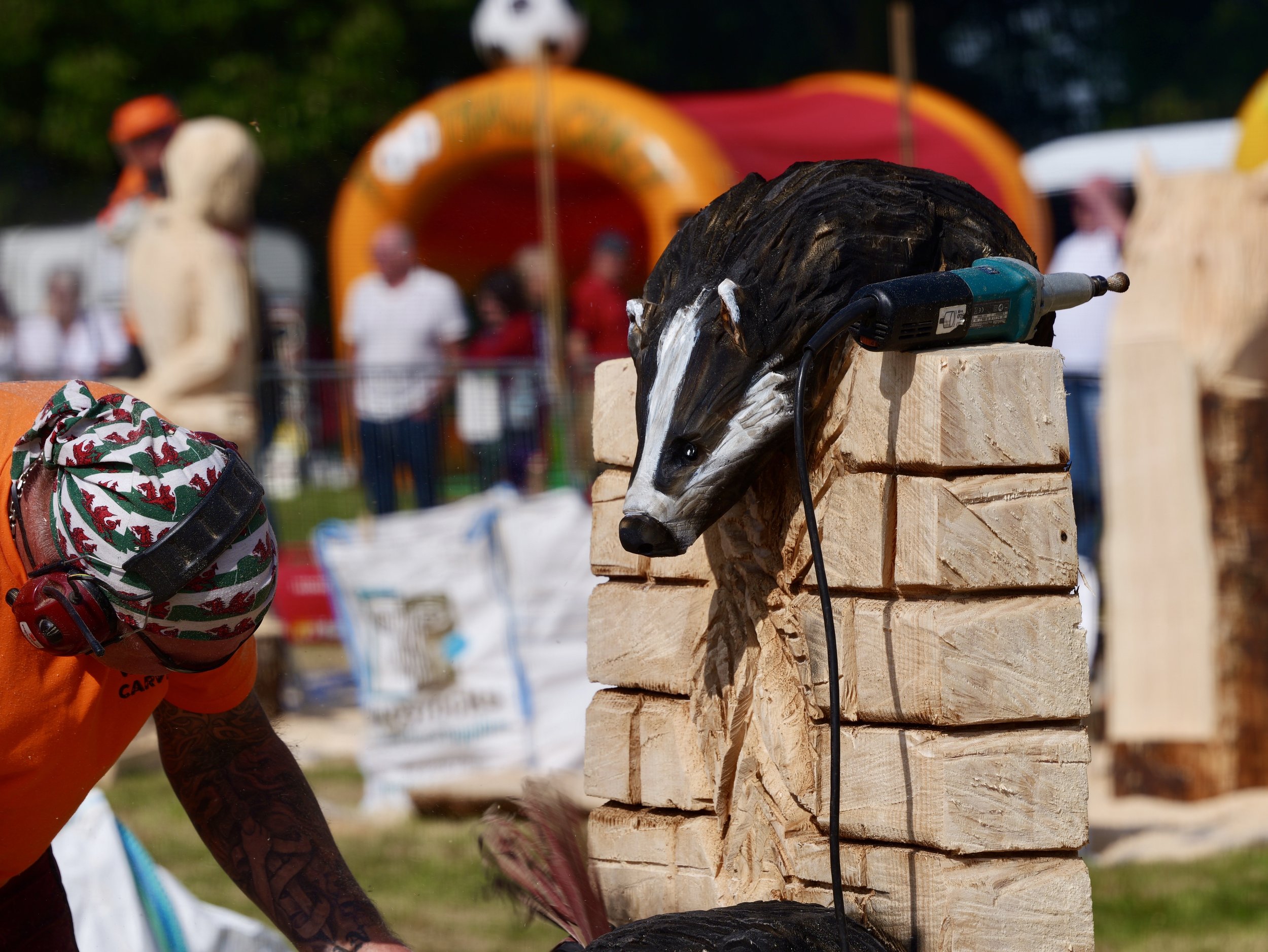 A worker in an orange shirt and holiday-themed hat carving a wooden sculpture of a hedgehog, with a power tool on the sculpture, at an outdoor event with people and colorful tents in the background.