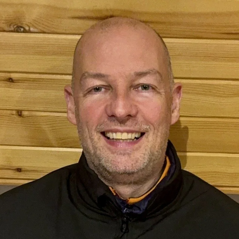 A smiling, bald man with a beard, wearing a black jacket, standing in front of a wooden wall.