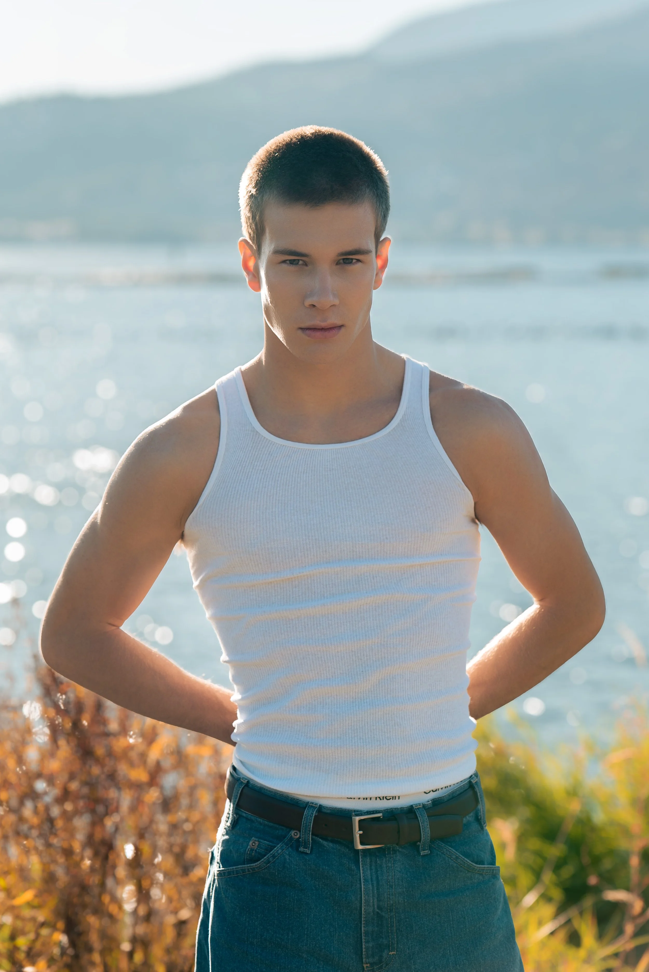 Young man wearing a white tank top and jeans standing outdoors near water with mountains in the background.