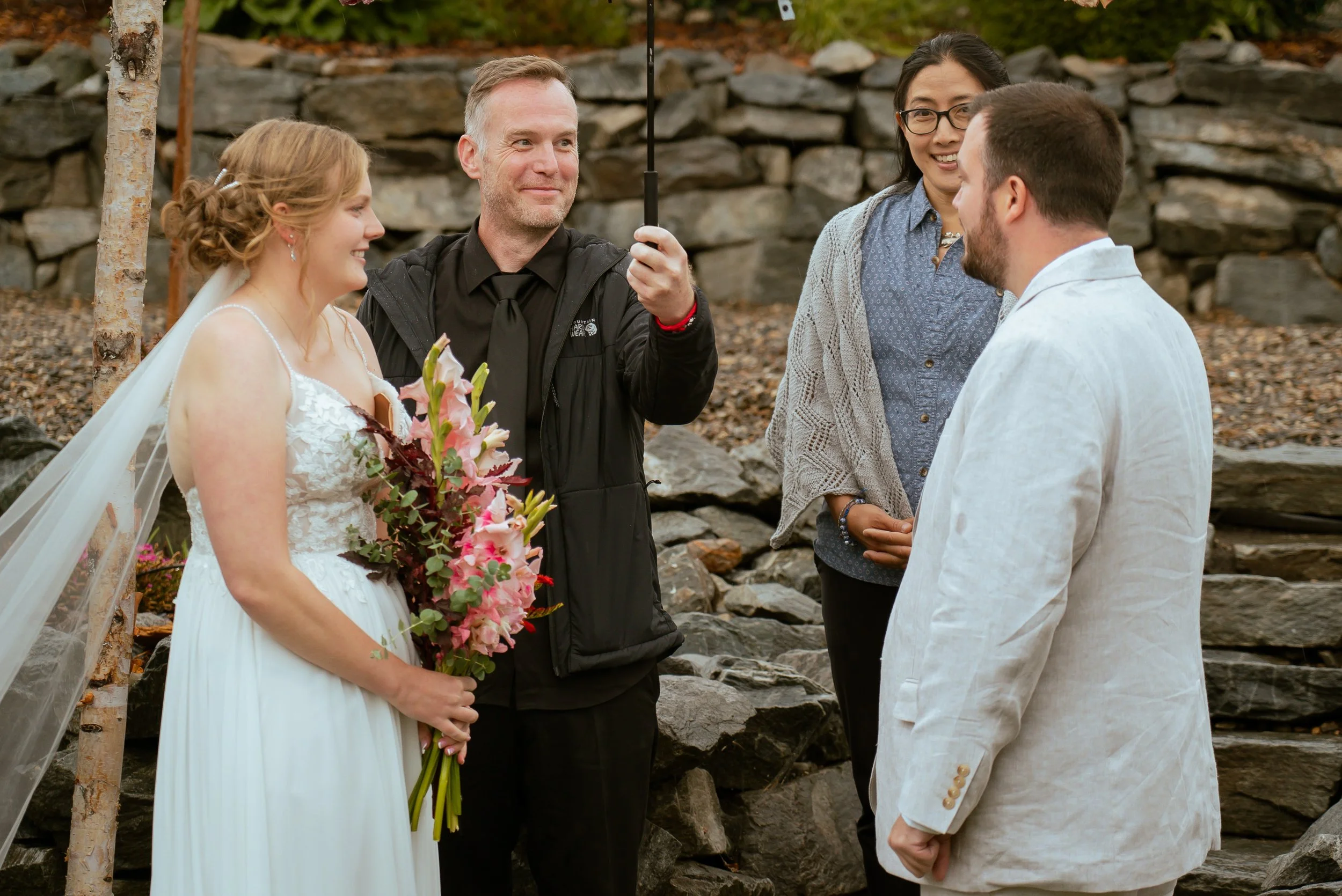 A wedding ceremony taking place outdoors with a bride, groom, officiant, and a woman standing with them, shot during the day near rocky terrain.