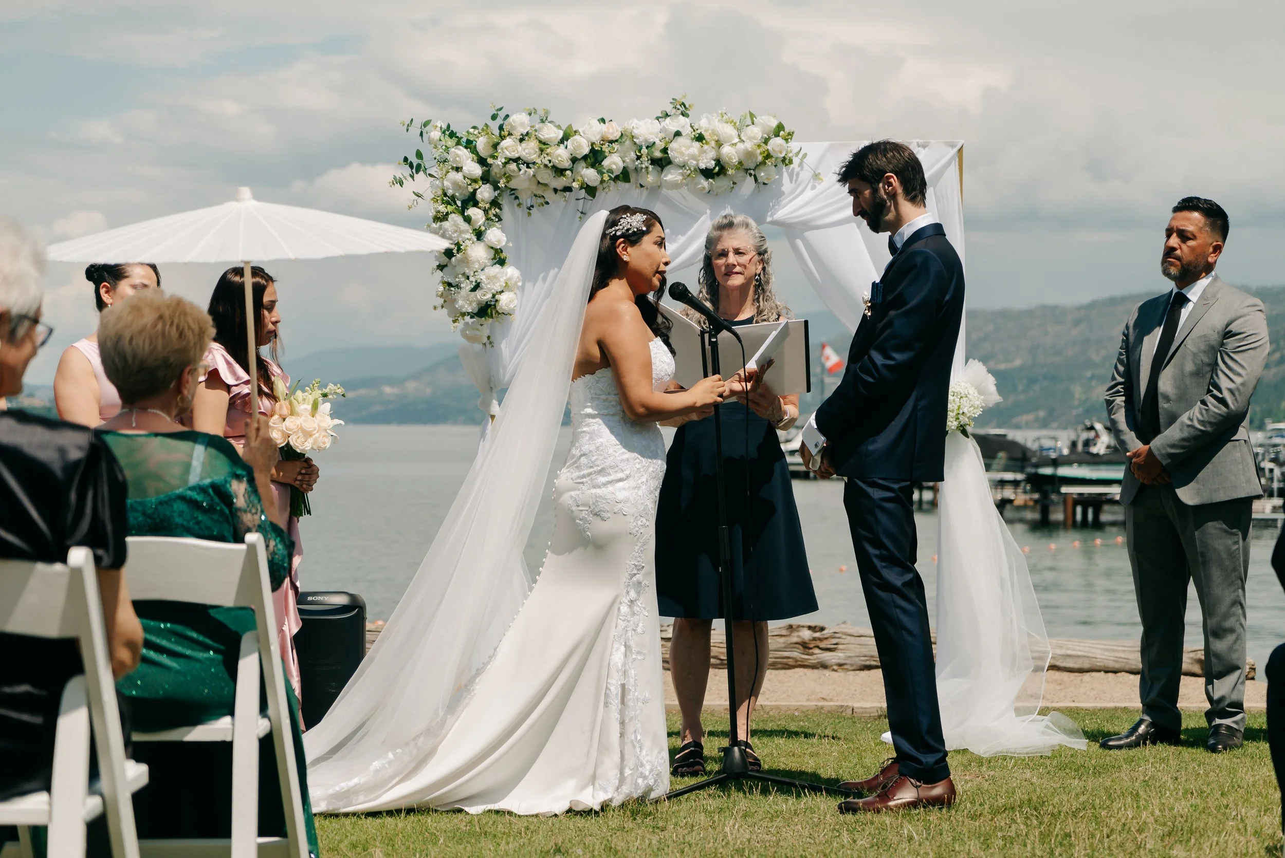 A wedding ceremony taking place outdoors by a lake with mountains in the background, featuring a bride in a white gown and a groom in a navy suit, standing under a white floral arch with an officiant reading from a book.