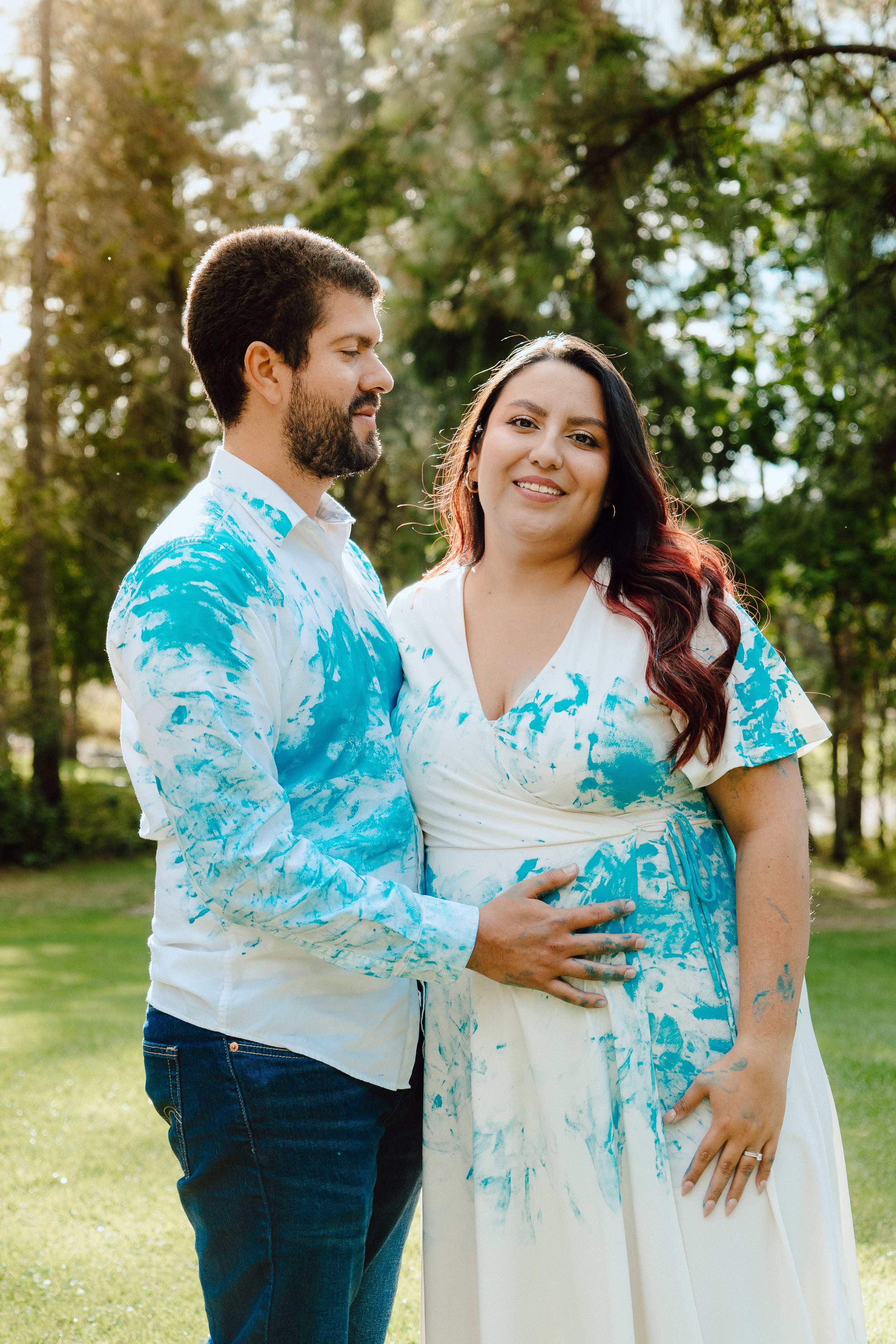 A couple stands outdoors on a grassy area with trees in the background, both wearing white outfits splattered with blue paint. The man is holding the woman's belly, and they are smiling at the camera, celebrating pregnancy.